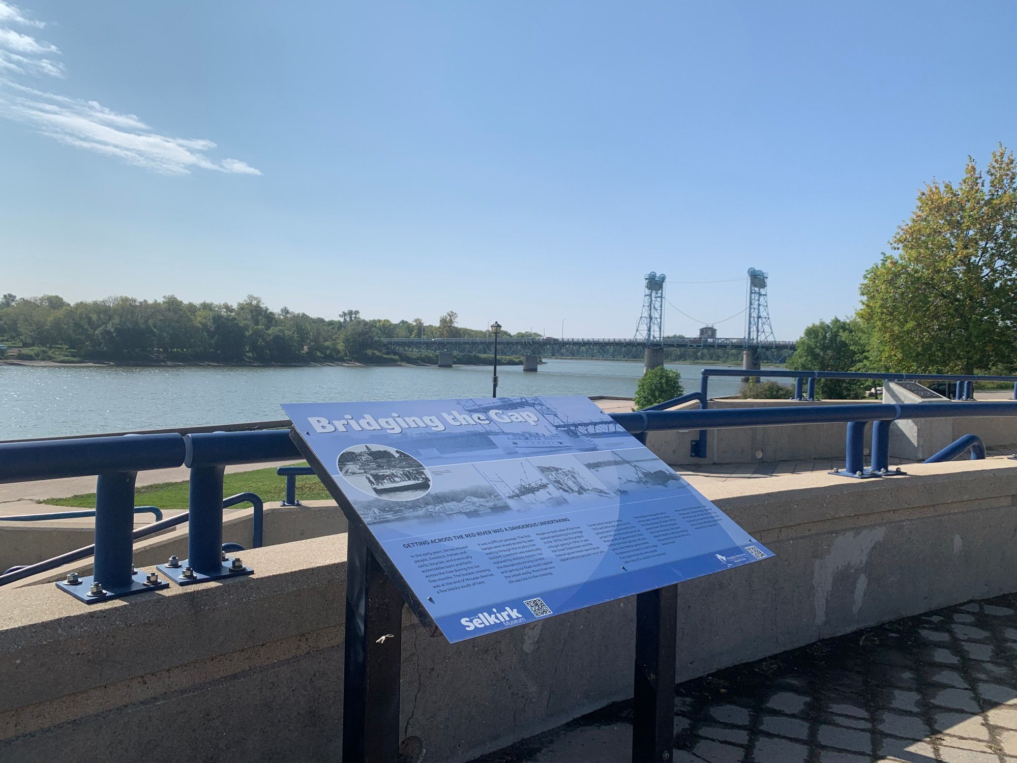 A blue interpretive sign mounted along the Red River in Selkirk Manitoba. It tells the story of the construction of the lift bridge that can be seen in the distance crossing the river. It's a sunny, summer day and all of the trees along the riverbank are green.