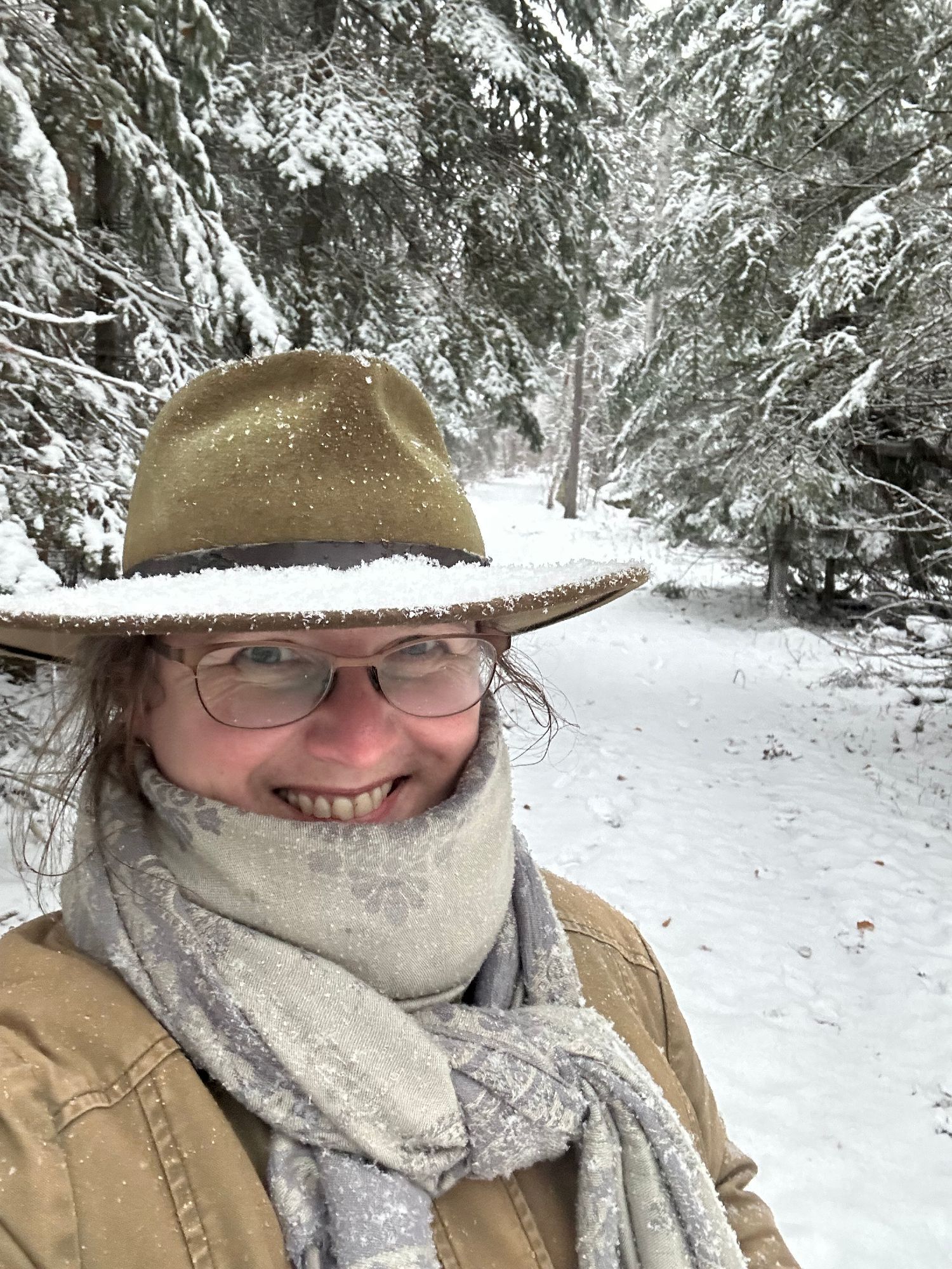 A selfie of a woman wearing a brown jacket and wide-brimmed felt hat, which is covered in fresh snow. She's smiling at the camera from behind a light grey scarf bundled around her heck. Behind her is a snow-covered spruce forest.