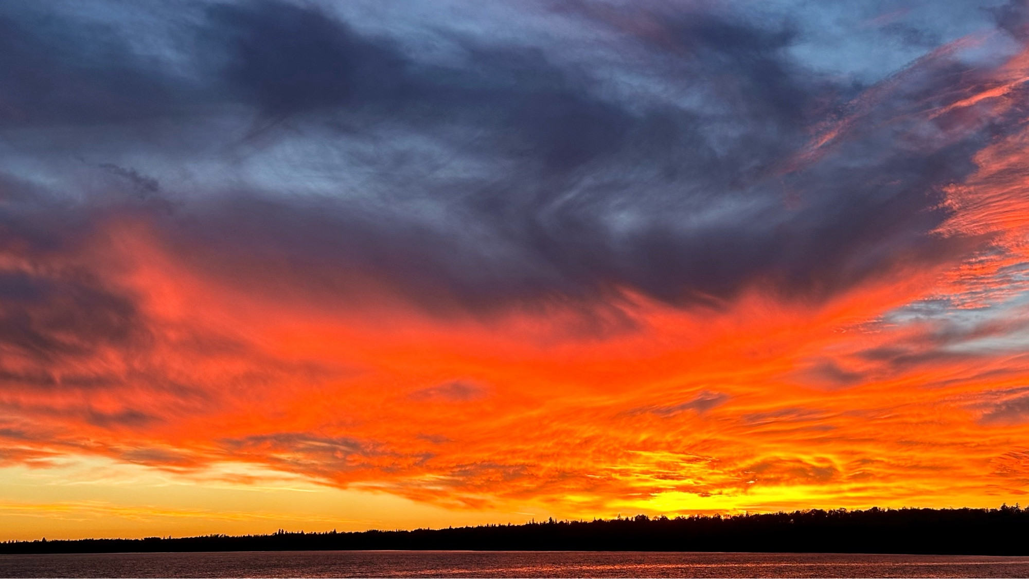 The sky looks like it’s on fire right above the horizon and dark red, flares up into the underside of wispy gray clouds that reach up to the top of the photo. in front of this dramatic sunset is a line of dark trees weigh in the distance bounding the shore of a very large lake.