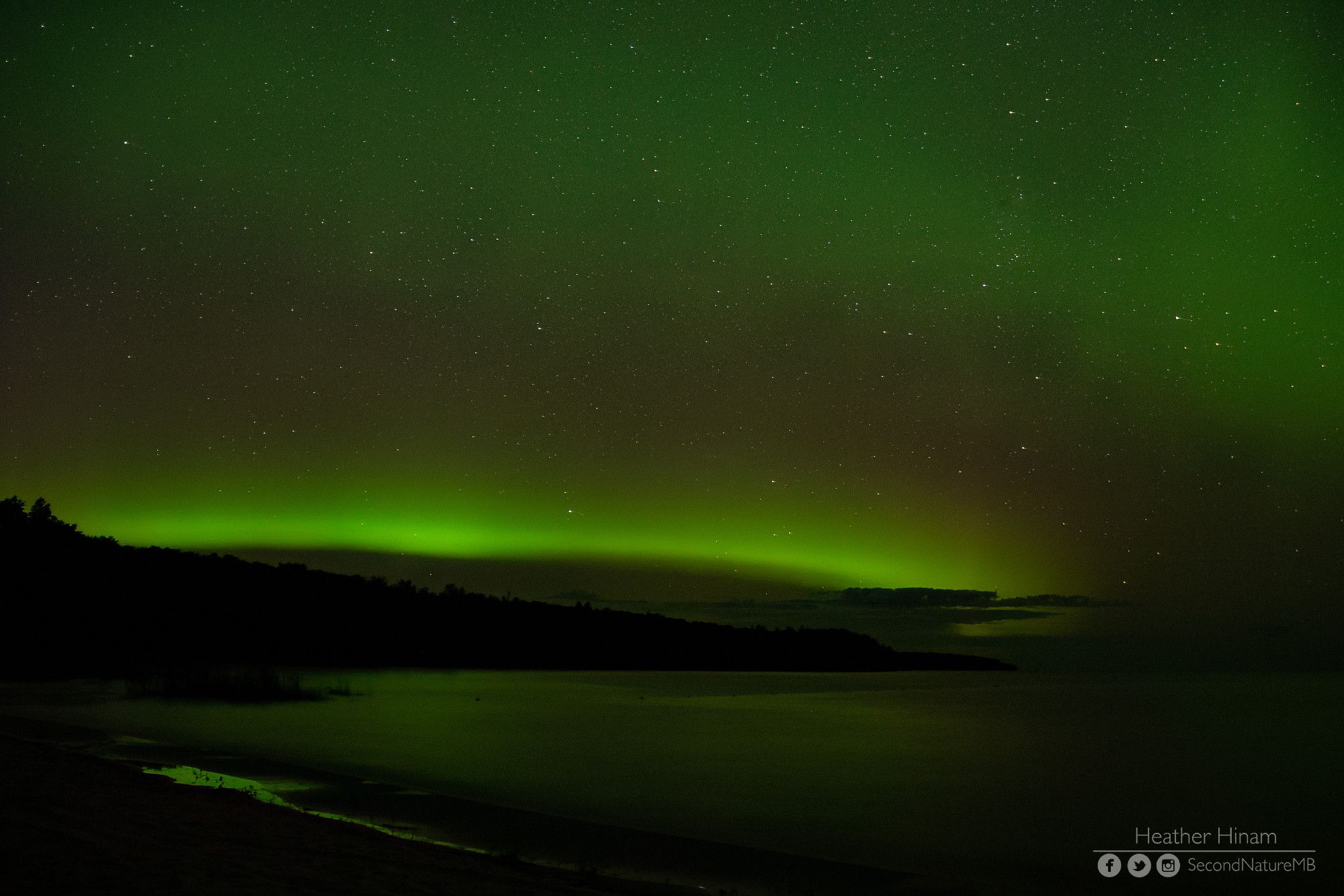 A band of bright green aurora arcs over the shoreline of Lake Winnipeg. The sky above it is a bit red and then more green fills the rest of the night sky.