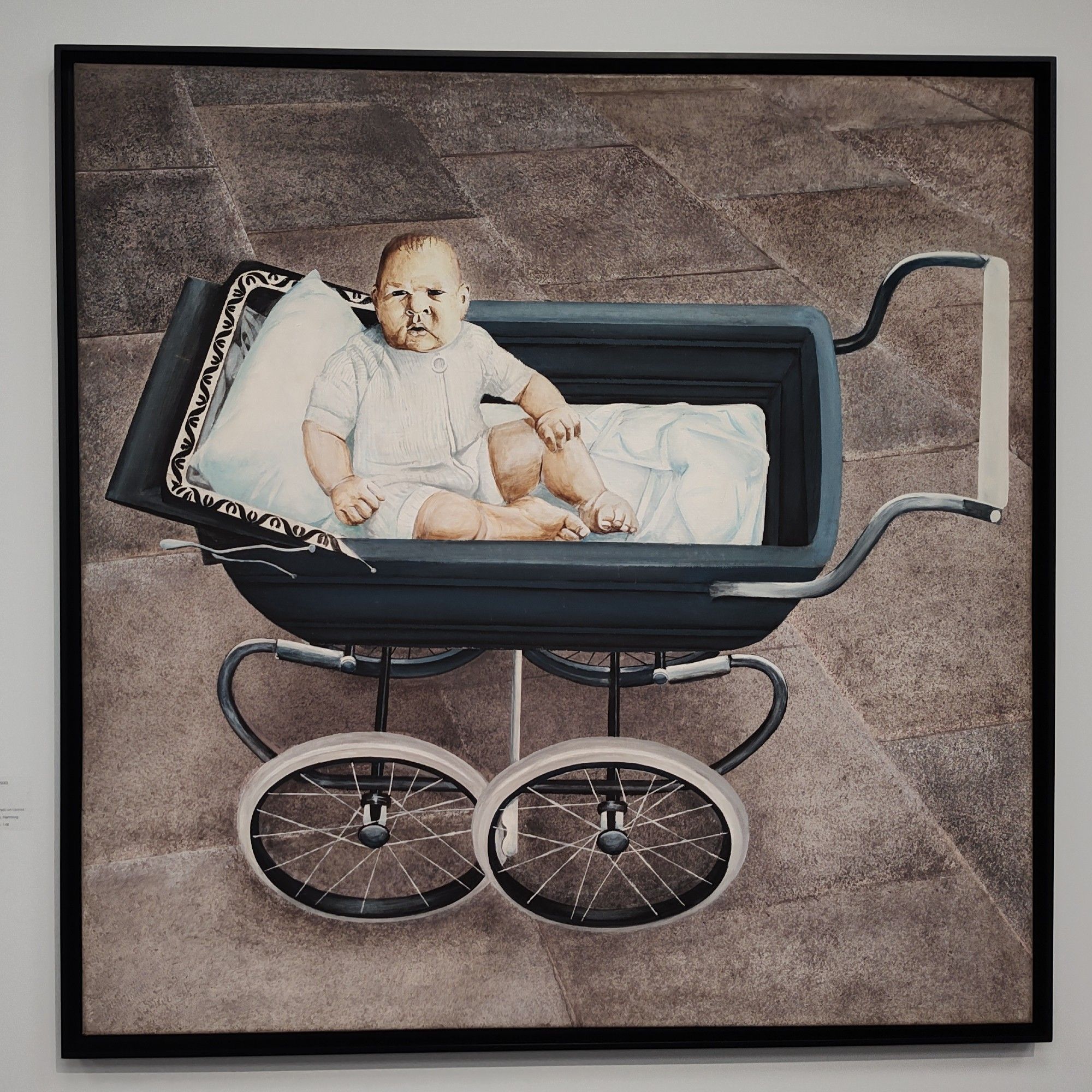 Das Gemälde "Das Wunschkind" von Hans Platschek, 1972, Acrylgemälde eines ernst und skeptisch schauenden, in einem grauen Kinderwagen mit großen Rädern sitzenden Babys in weißem Oberteil, Kinderwagen auf grau-beigen Gehwegplatten, fast photorealistisch gemalt aber von den Dimensionen und Perspektiven irritierend verfremdet