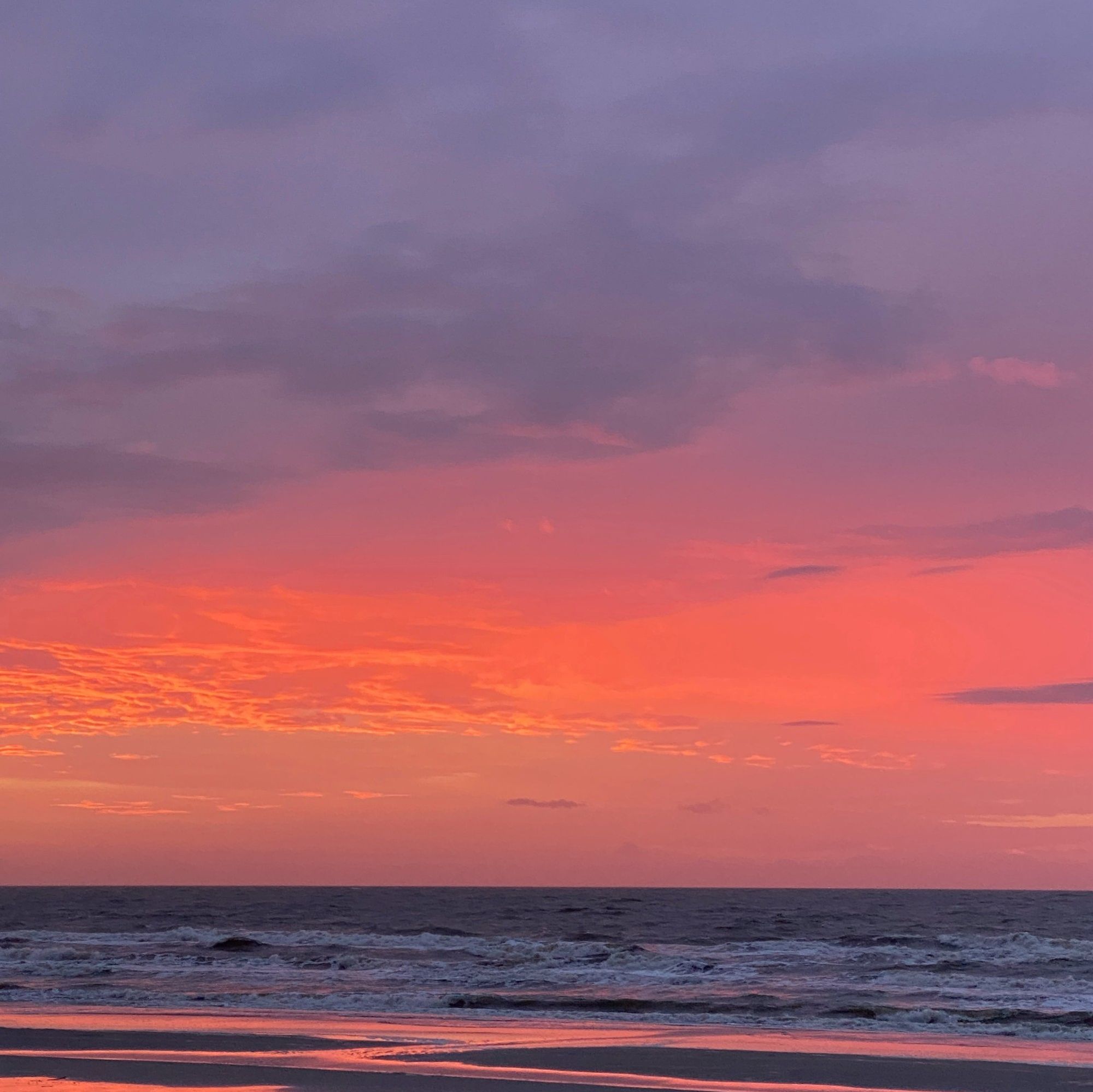 The sun rises over the ocean coloring the sky in orange, violet, pink, and blue. The colors are reflecting on the water.