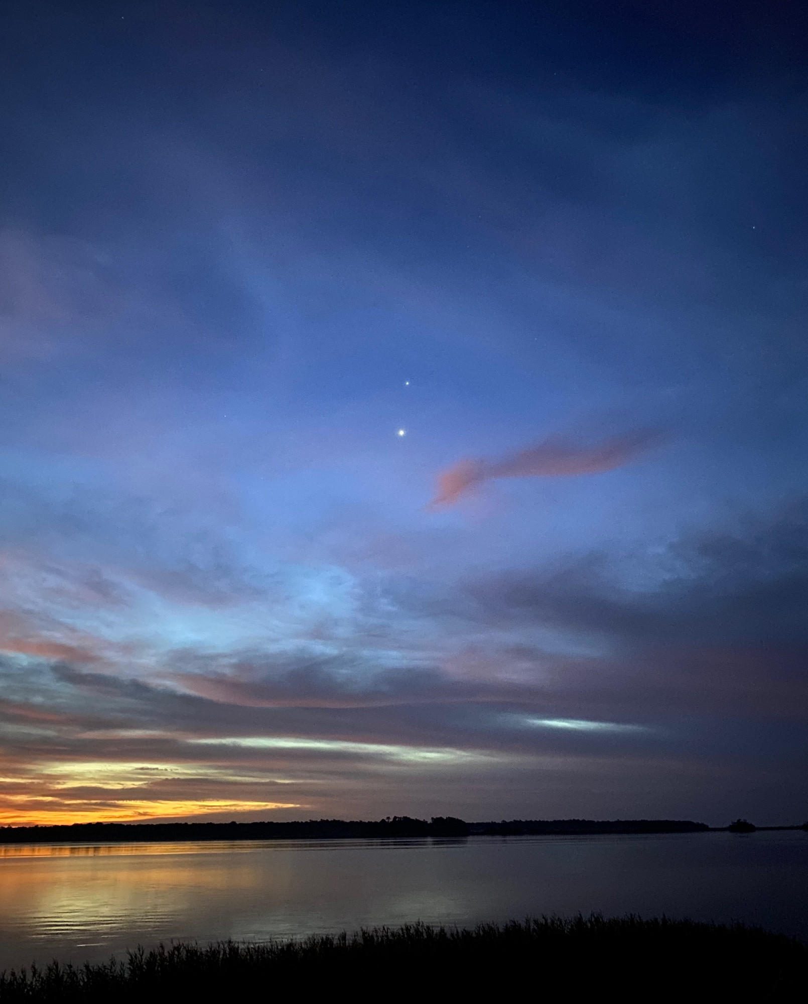 Sunshine is peeking through clouds on the left and the night sky on the right. Below is still water showing the sky’s reflection. In the middle is Jupiter and Venus. 