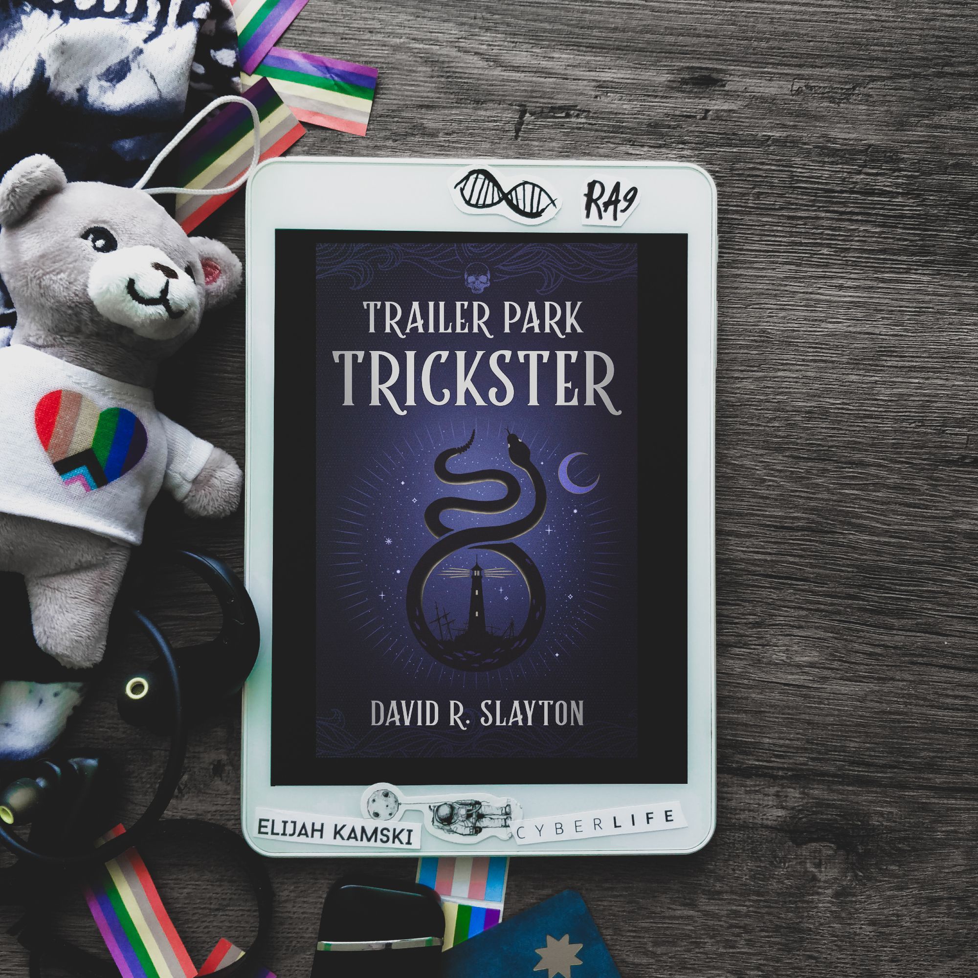 trailer park trickster by david r. slayton, second book in adam binder trilogy. purple cover with snake wound around a tower or a light house, with moo and stars around it. white framed tablet is placed on a dark grey wooden table, with various items shoved to the left side of it, such as skull-pattern mask, teddy bear with progress pride flag, and pride flags confetti.