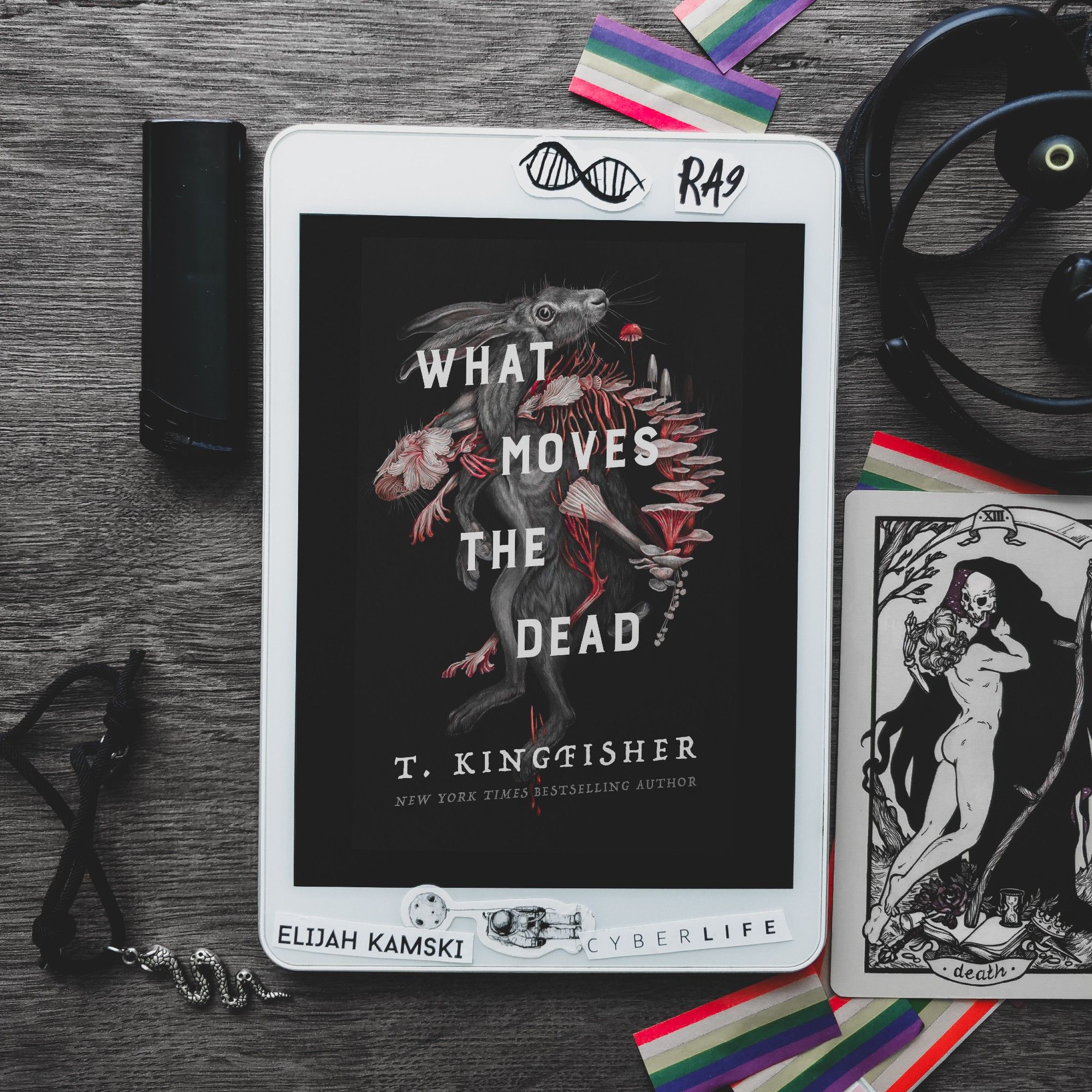 what moves the dead by t. kingfisher, first book in Sworn Soldier series. Black cover, with two rabbits or hares intertwined, one normal, the other one - dead, and made mostly of bones and fungi. White framed tablet is placed on a dark grey wooden table, with various items strewn around. such as a bracelet with snake charm, a tarot card with reaper kissing a person, earphones, black lighter, and pride flag confetti. Picture by Wannabe Kingpin, review by Reading Nosferatu