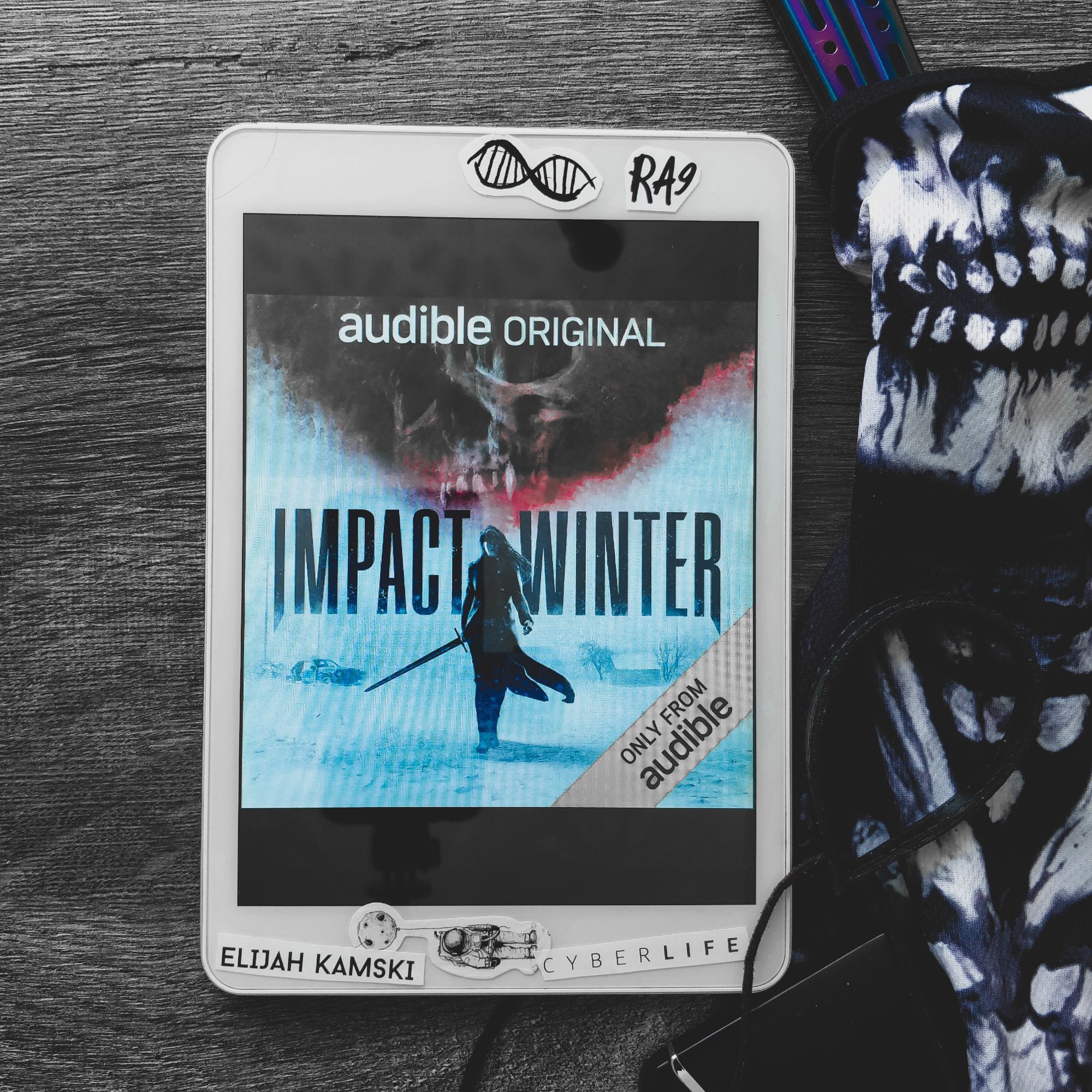 impact winter by travis beacham audiobook from audible original a warrior with a sword is walking through snowed up field, with sky tearng apart above them, showing a fanged skull. white-framed tablet is placed on a dark grey wooden table with skull face facemask beside it and a few other dark items