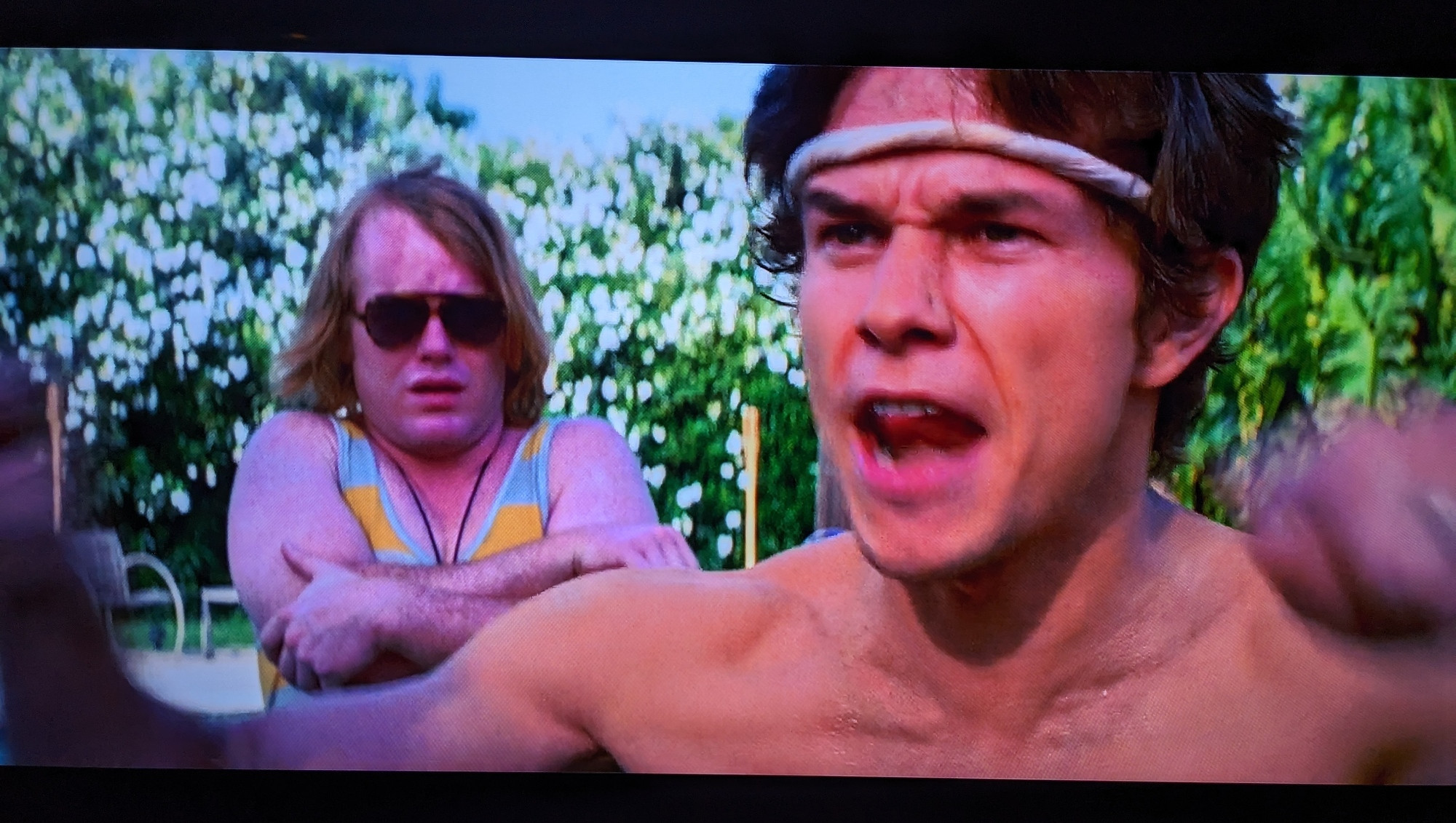 Scotty J looking horribly distressed and scared with his arms crossed standing behind Dirk Diggler while Dirk screams