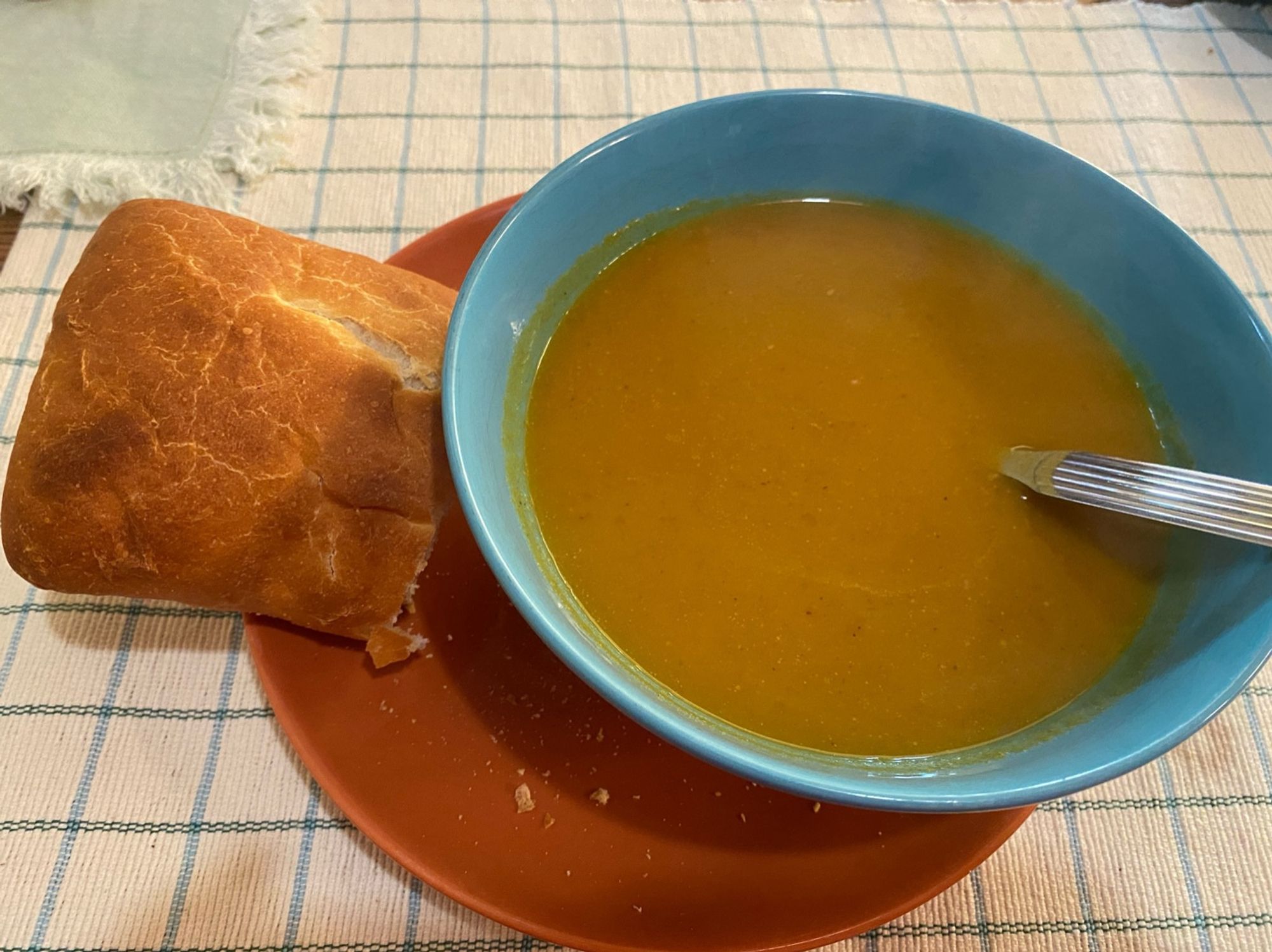 Lichtblauw kommetje met warmgele soep, op een oranje bordje met een stuk ciabatta 