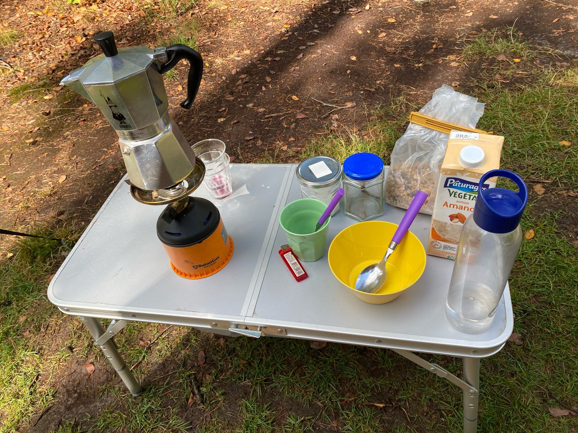 Campingtafeltje in het bos met erop een butaanbrandertje met pruttelpotje, twee lege wijnglazen, een aansteker, potje suiker, leeg potje waar koffie in zat, een groen campingbekertje met een paars lepeltje, een geel campingbakje met grote paarse lepel, een zak cruesli, een pak amandelmelk en een bijna leeg waterflesje