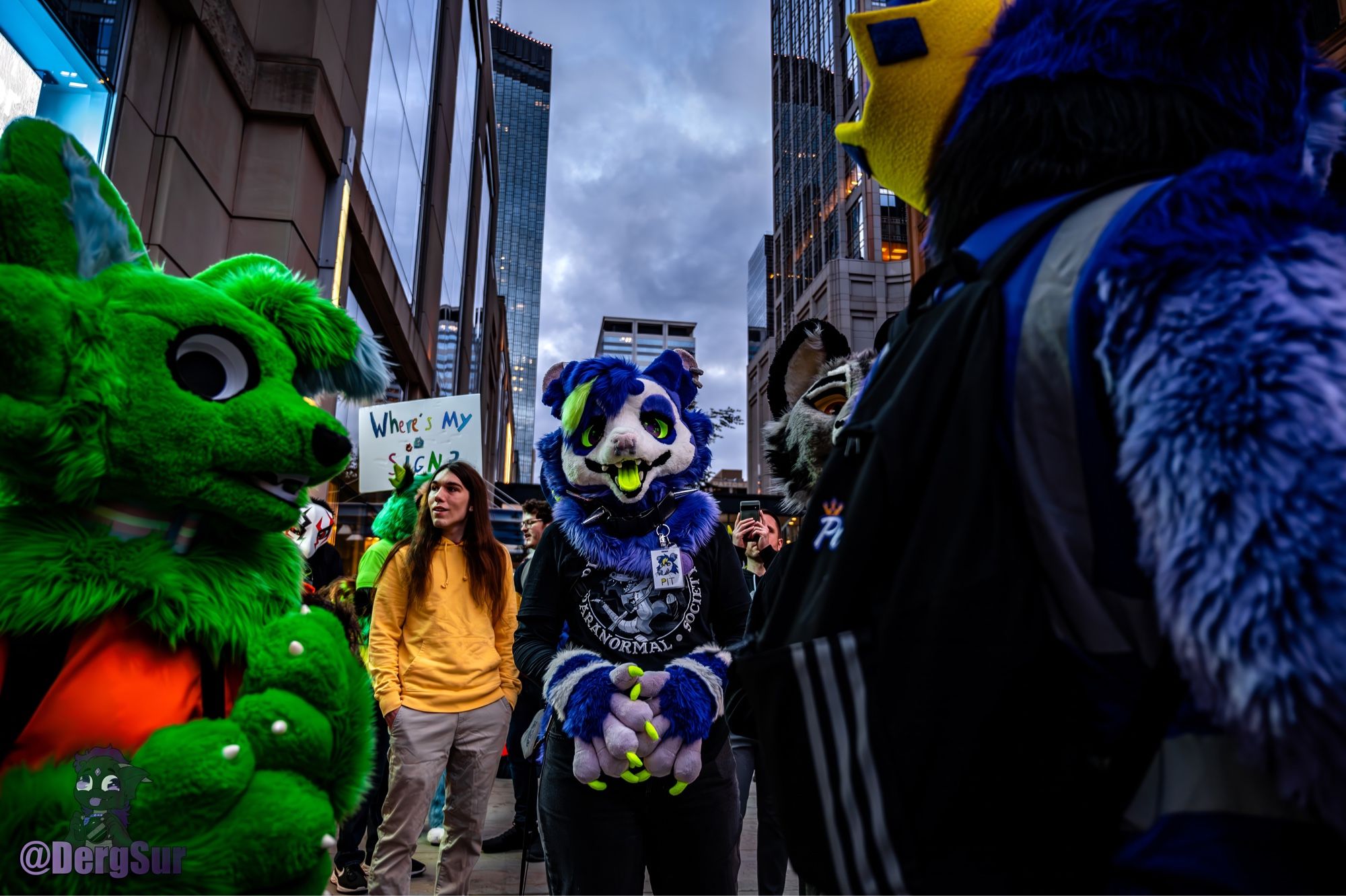 Purple possum fursuiter surrounded by other fursuiters with tall buildings in the background