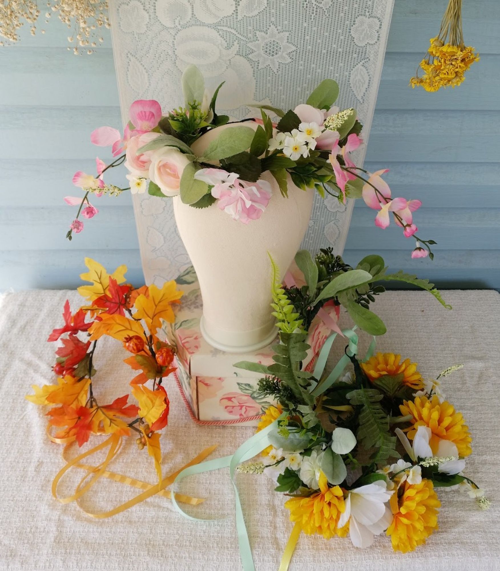 photograph of a mannequin head wearing a flower crown. several more flower crowns sit at the base of its stand. dried florals and a harvest-themed lace backdrop are in the background.