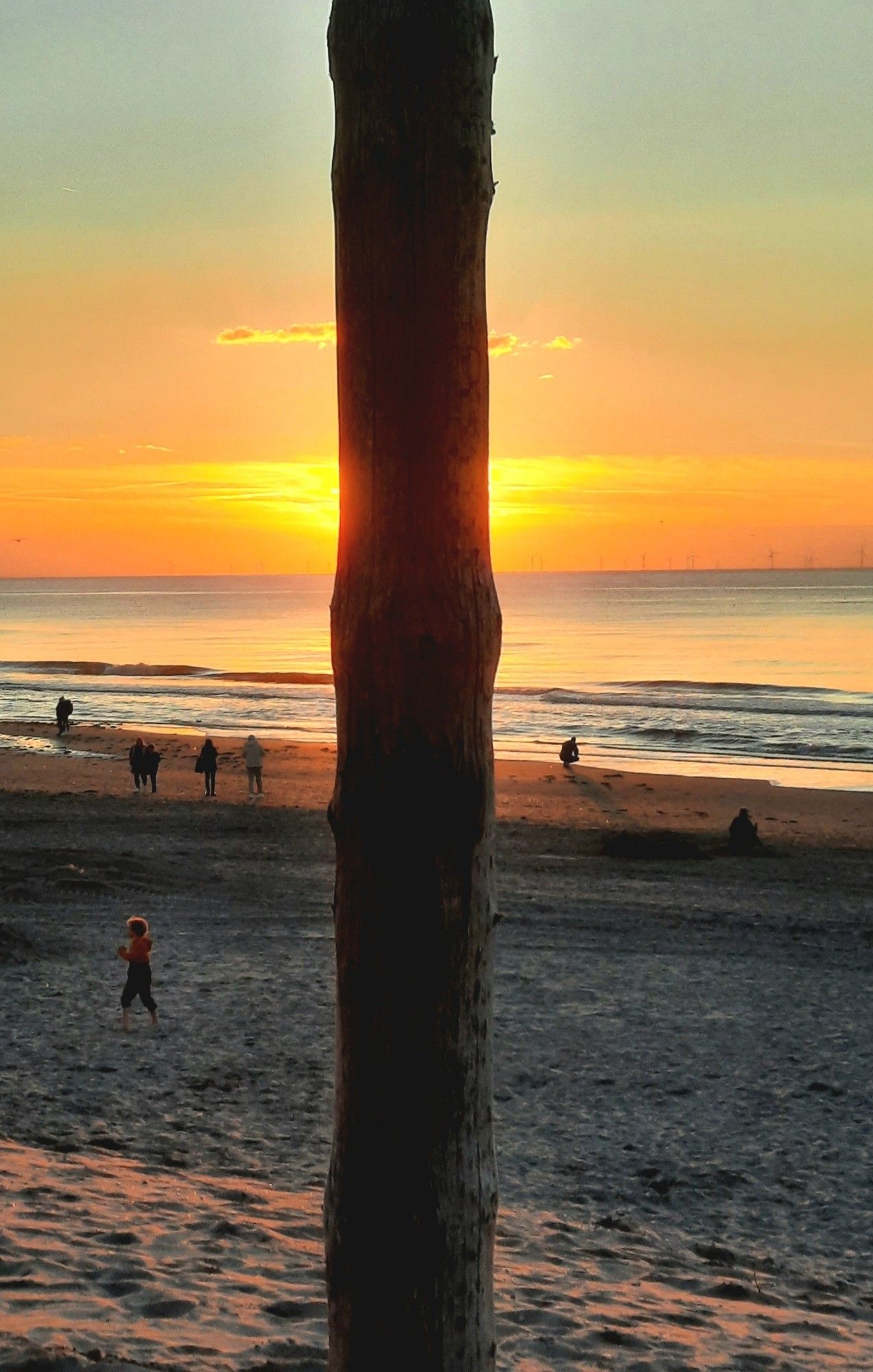 Orange gefärbter, fast wolkenloser Himmel über der ruhigen Nordsee. Von der untergehenden Sonne ist nichts zu sehen, da ein 20 Zentimeter breiter senkrechter Holzmast eines Strandpavillons die Sicht versperrt. Nur genau dahinter glüht der Himmel.