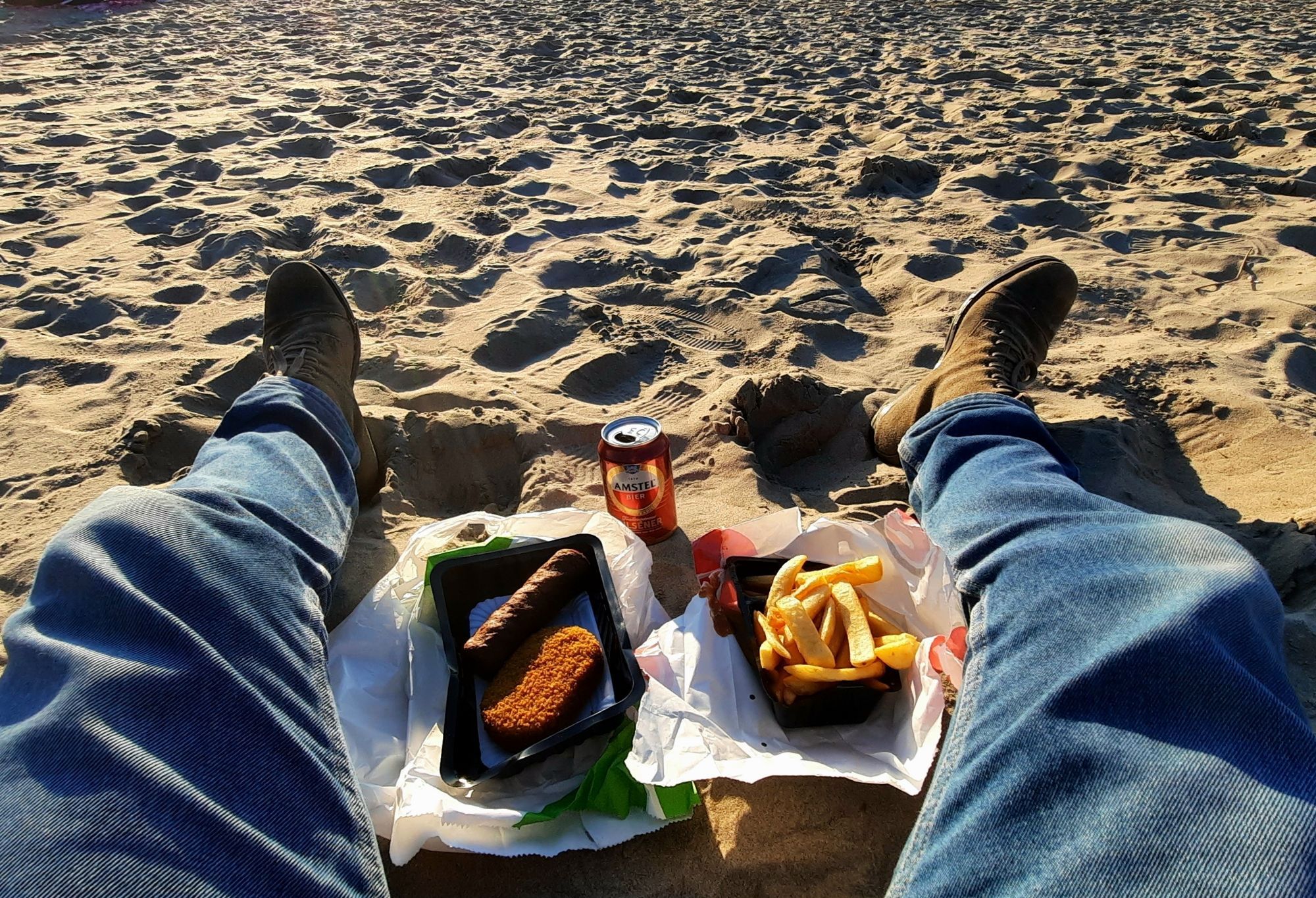 Ich sitze mit blauer Jeans und Boots im Sand. Zwischen meinen Beinen eine Pommes, eine Schale mit 2 Snacks und eine rote Dose Amstelbier.