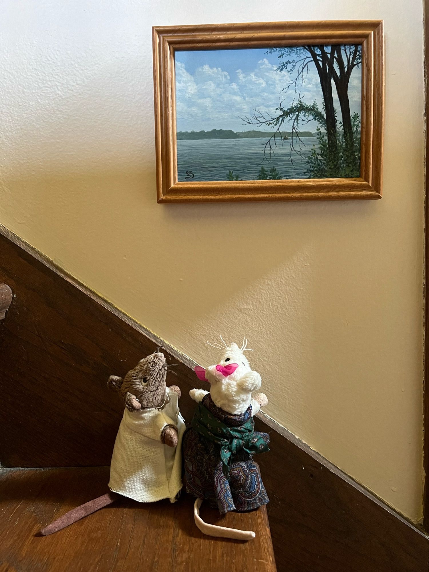 Two toy mice stand on a wooden step looking at a small landscape painting hung just above them in the stairwell.