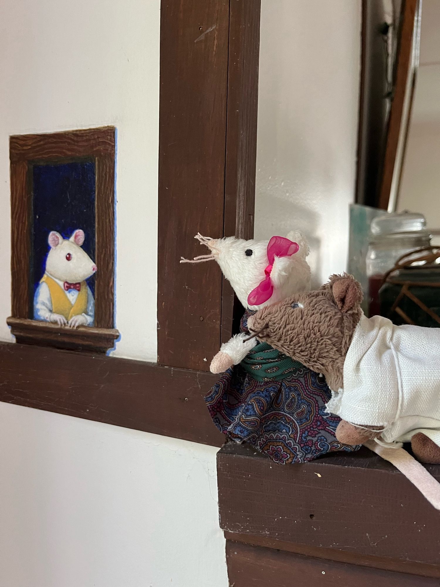 Two toy mice on the end of a mantlepiece looking at a window painted on the wall beside them. In the window a painted mouse the same size looks back.