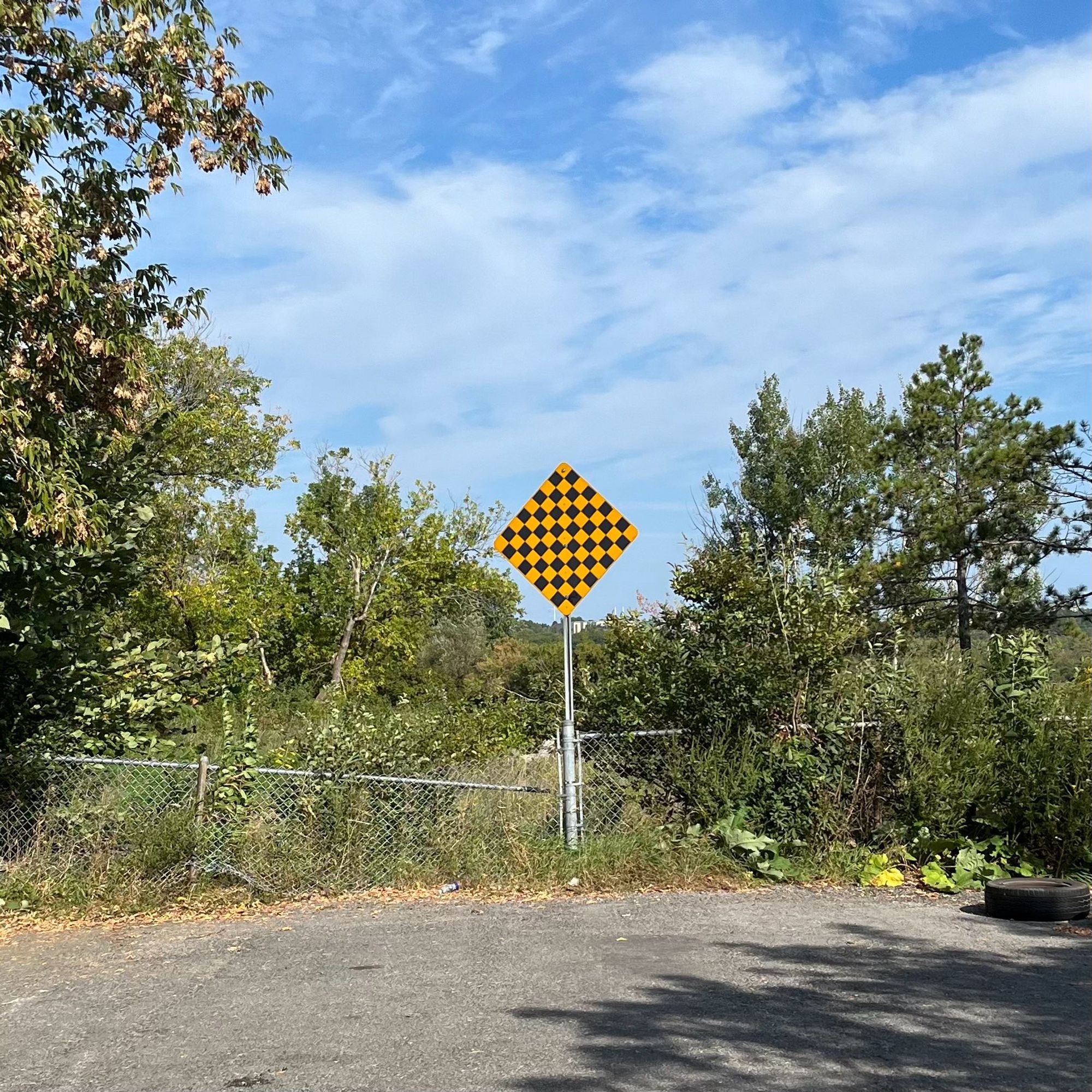 Photograph of the end of a road. The tarmac stops at an overgrown and broken chainlink fence. In the centre is a large diamond-shaped warning sign in yellow and black lozenges.  Beyond the sign and fence is scrub and meadow with distant hills in the background.