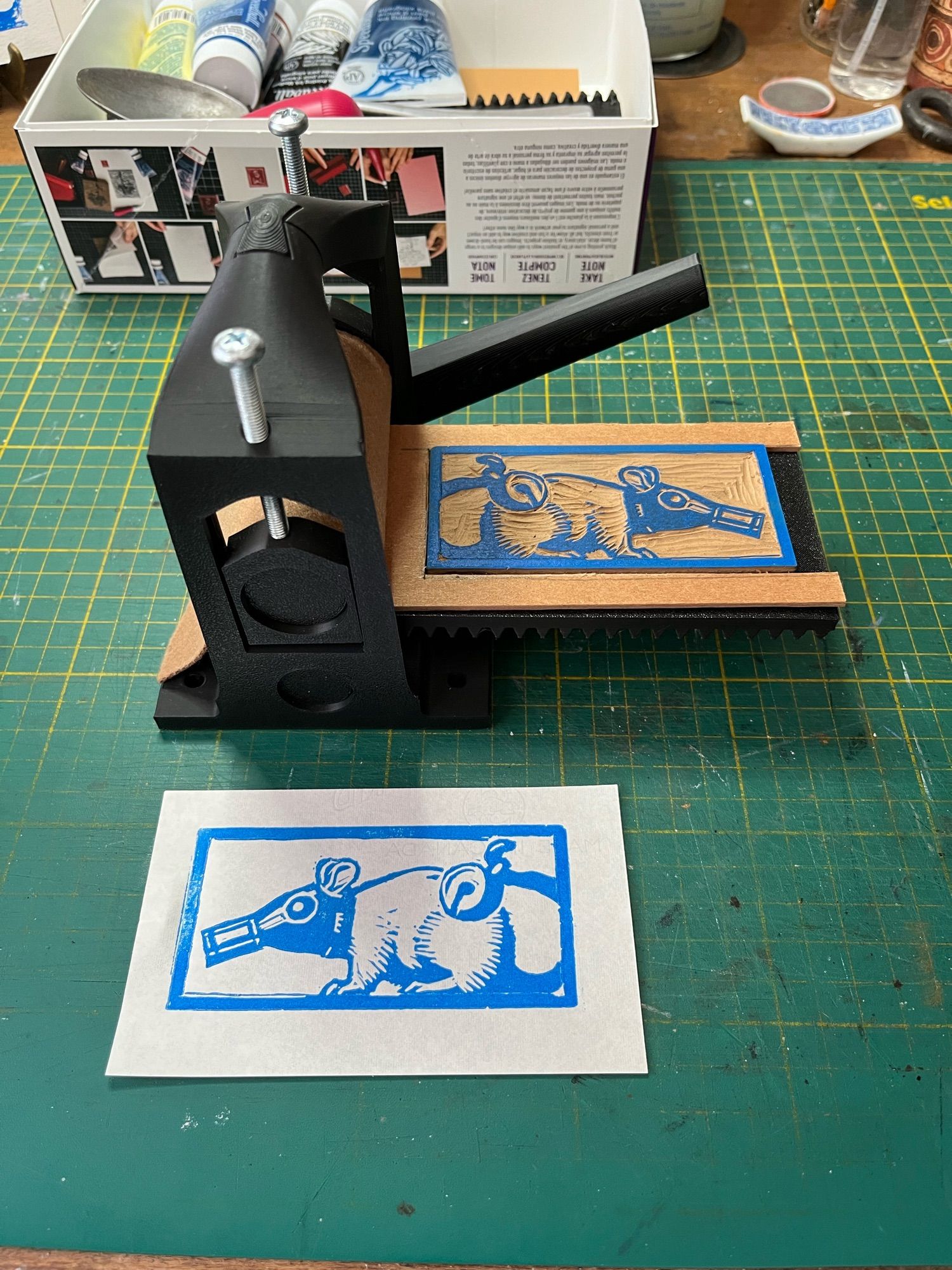 Photograph of a small plastic printing press with a carved piece of linoleum on the bed. A print from that block lies on the table in front of the press, showing an image of a rat wearing a gas mask and holding a bell on its tail.