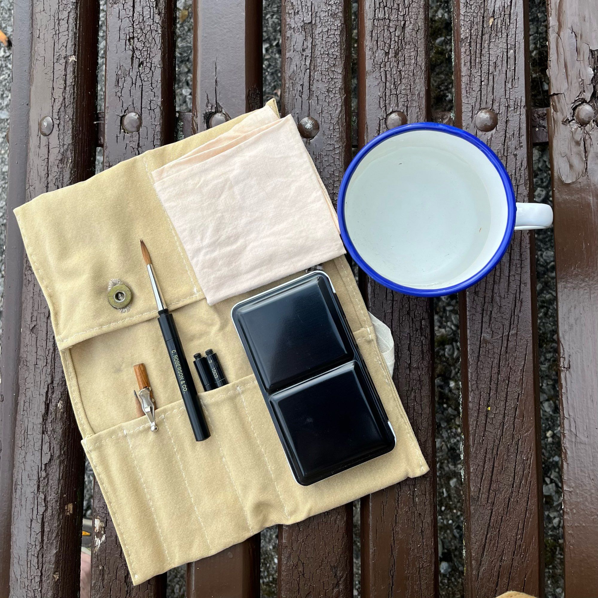 Photograph of a portable watercolour painting set and an enamelled mug of water on a park bench