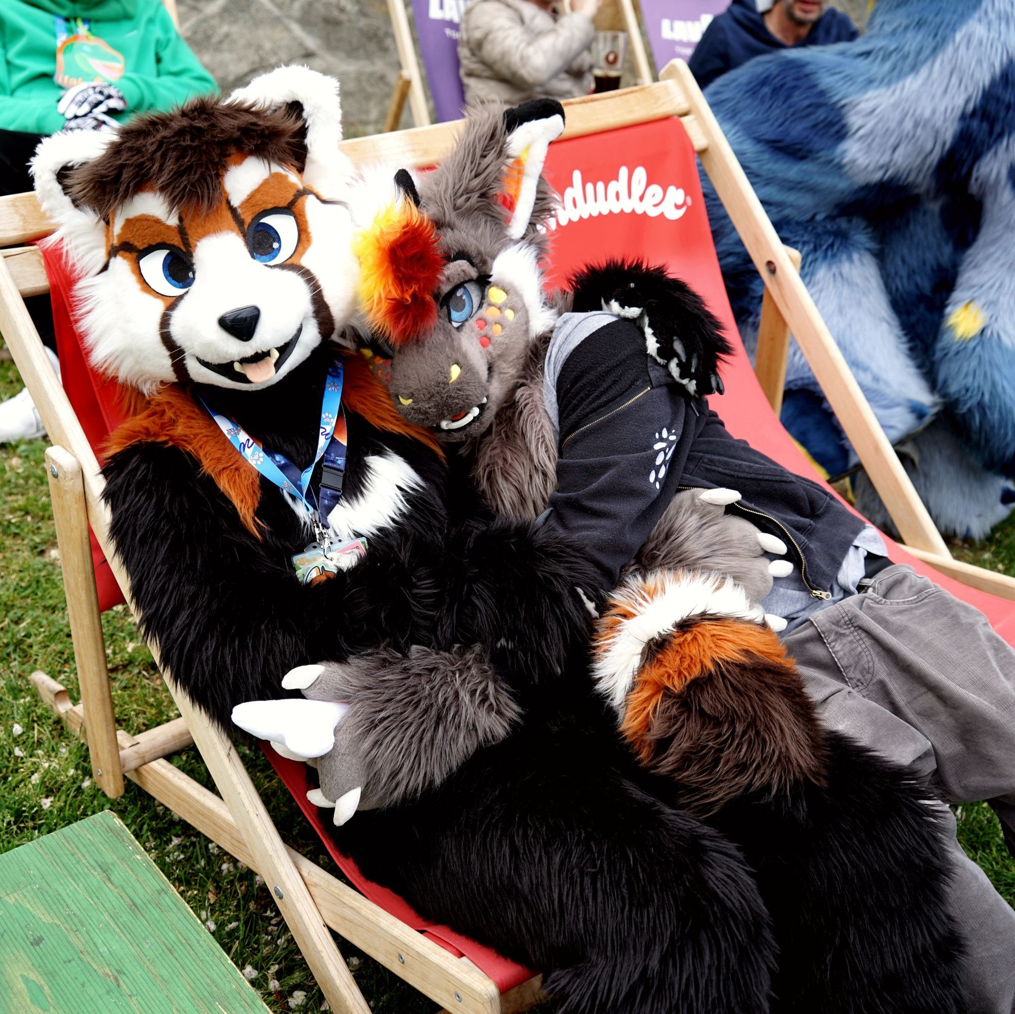 A red panda and a dragon fursuiter cuddling on two folding chairs in a garden