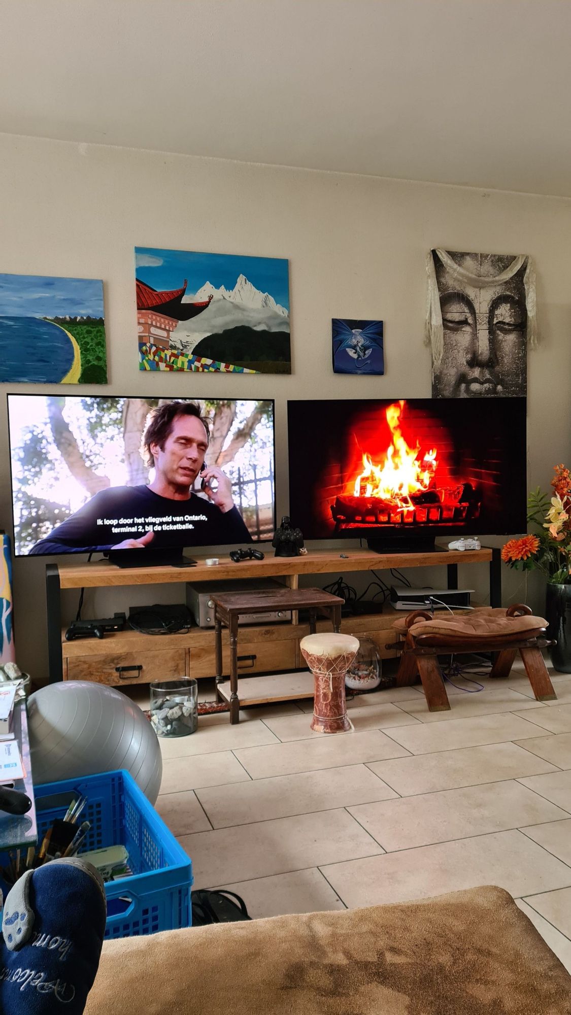 Foto van een muur met 3 schilderijen (zee, Tibetaans klooster, Boeddha) en daaronder 2 tvs, links met een still uit de serie Prison Break en rechts met een haardvuurtje op het scherm.