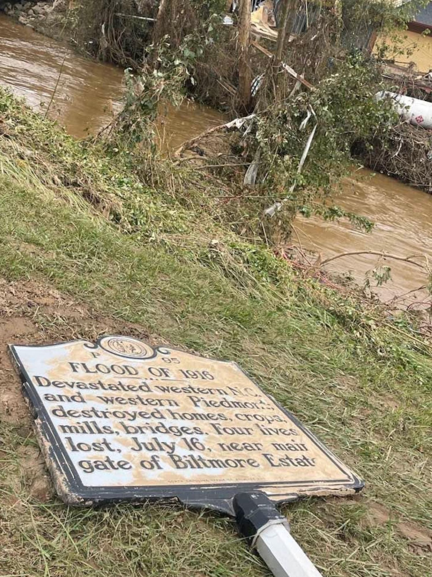 an historical marker for the Asheville Flood of 1916, flat on the ground in the grass beside muddy flood waters