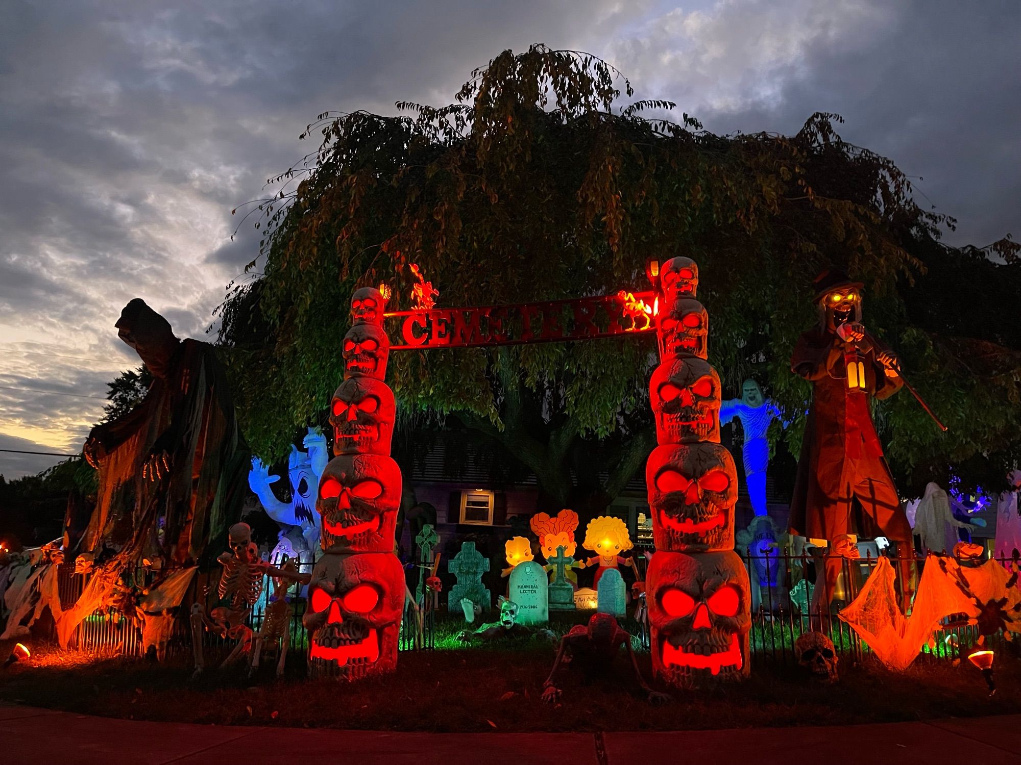 A series of Halloween decorations are backlit by the remains of the day. There are gravestones, ghouls, ghosts, and ghastlies with lit up eyes and various implements.