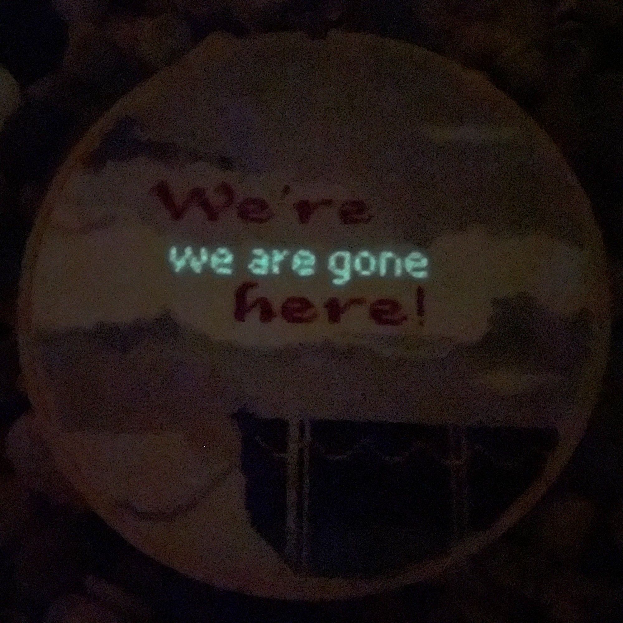 A photo taken in the dark of the same embroidery, but hidden between the two lines that read 'we're here!' are the words 'we are gone' showing up in glow in the dark stitched thread