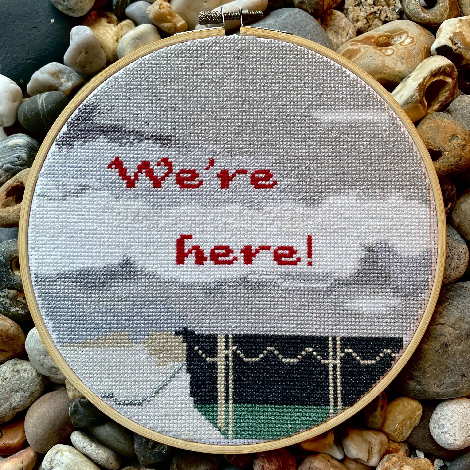 Cross stitch embroidery hoop showing a rowing boat on shale, with a huge cloudy sky above. The words 'we're here!' are stitched above in red thread