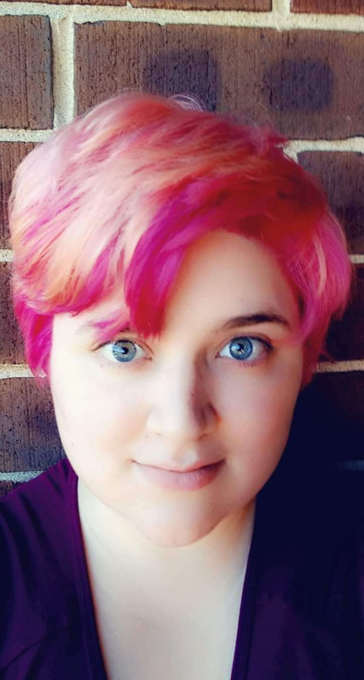 A person with short, vibrant pink and pale orange hair with gray eyes is smiling at the camera with a brick wall behind them.