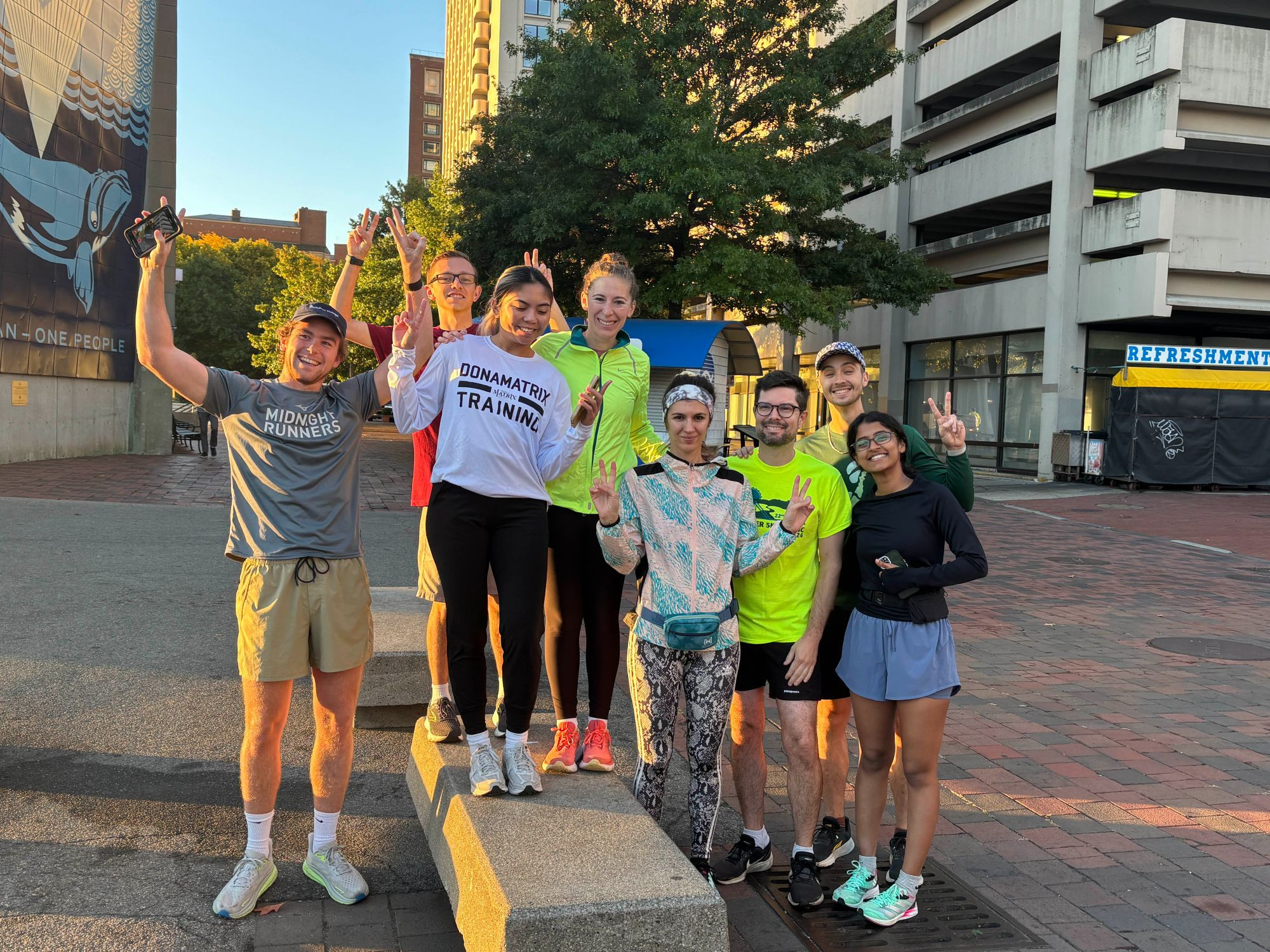 A group of enthusiastic runners poses for a post-run photo in the early morning sunlight, showing peace signs and smiles. They are standing in front of a city mural and an urban landscape, with the "Midnight Runners" logo visible on one participant's shirt. Everyone is dressed in bright, athletic gear, ready for the day's adventures!