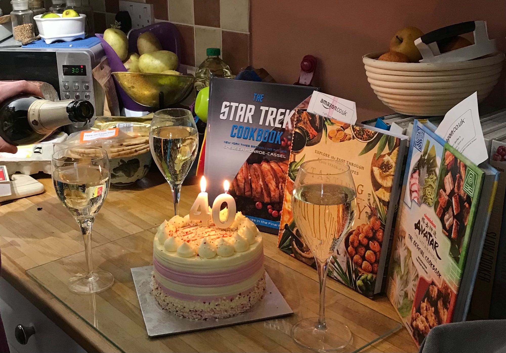 A display of the raspberry ripple birthday cake with lit 40 candles (the number shapes, not 40 separate candles), 3 glasses of prosecco, one still being poured, framed by the cook books i recieved: The Star Trek Cookbook, Tasting History by Max Miller & The Avatar the Last Air bender cookbook