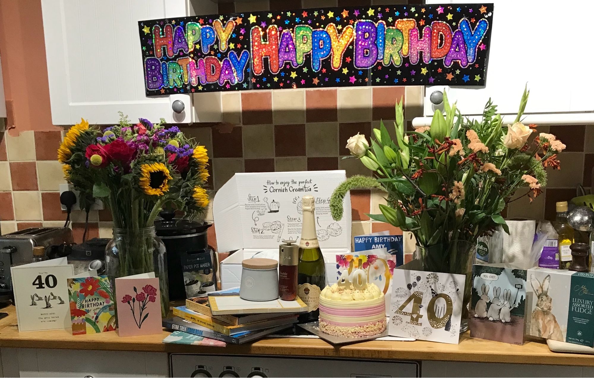 A spread of my birthday gifts in the kitchen under a happy birthday banner across the hob hood:  2 vases of stunning flowers, birthday cake, prosecco, birthday cards, a cornish cream tea, cook books, history books on pompeii, clarins serum, candles, fudge, chocolate & other lovely smol goodies!