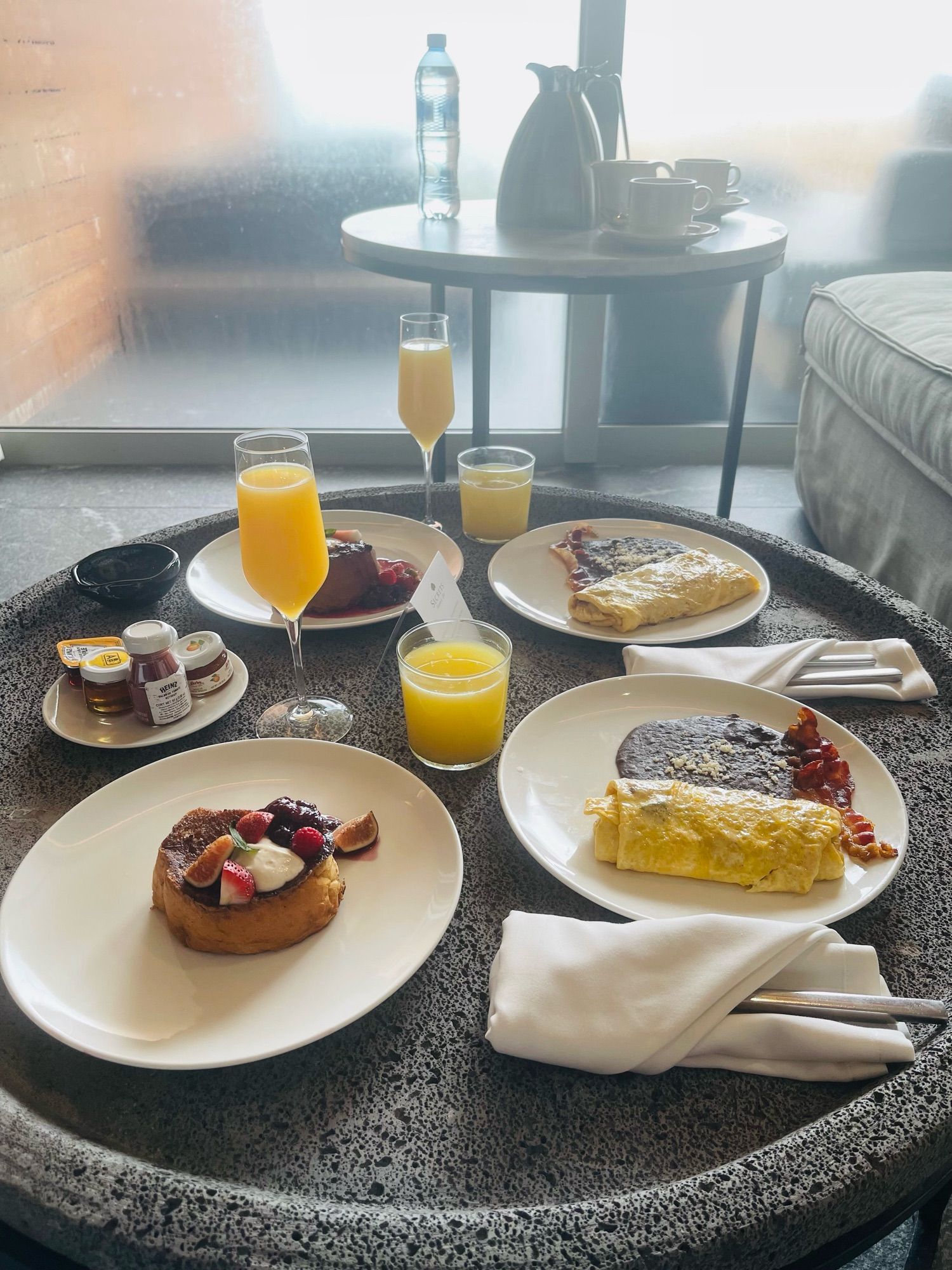 2 omelettes with refried black beans and bacon, and 2 massive pieces of french toast w/ berries and fresh figs on a gray stone coffee table arrayed with juices and condiments. A foggy sliding glass door is in the background