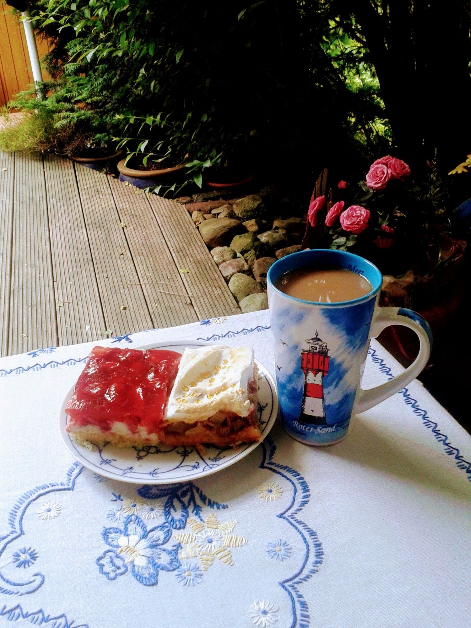 Großer Kaffeebecher und ein Teller mit 2 St Kuchen, stehen auf einem Gartentisch mit bestickter Tischdecke. Dahinter ist eine Holzterrasse und Pflanzen zu sehen.