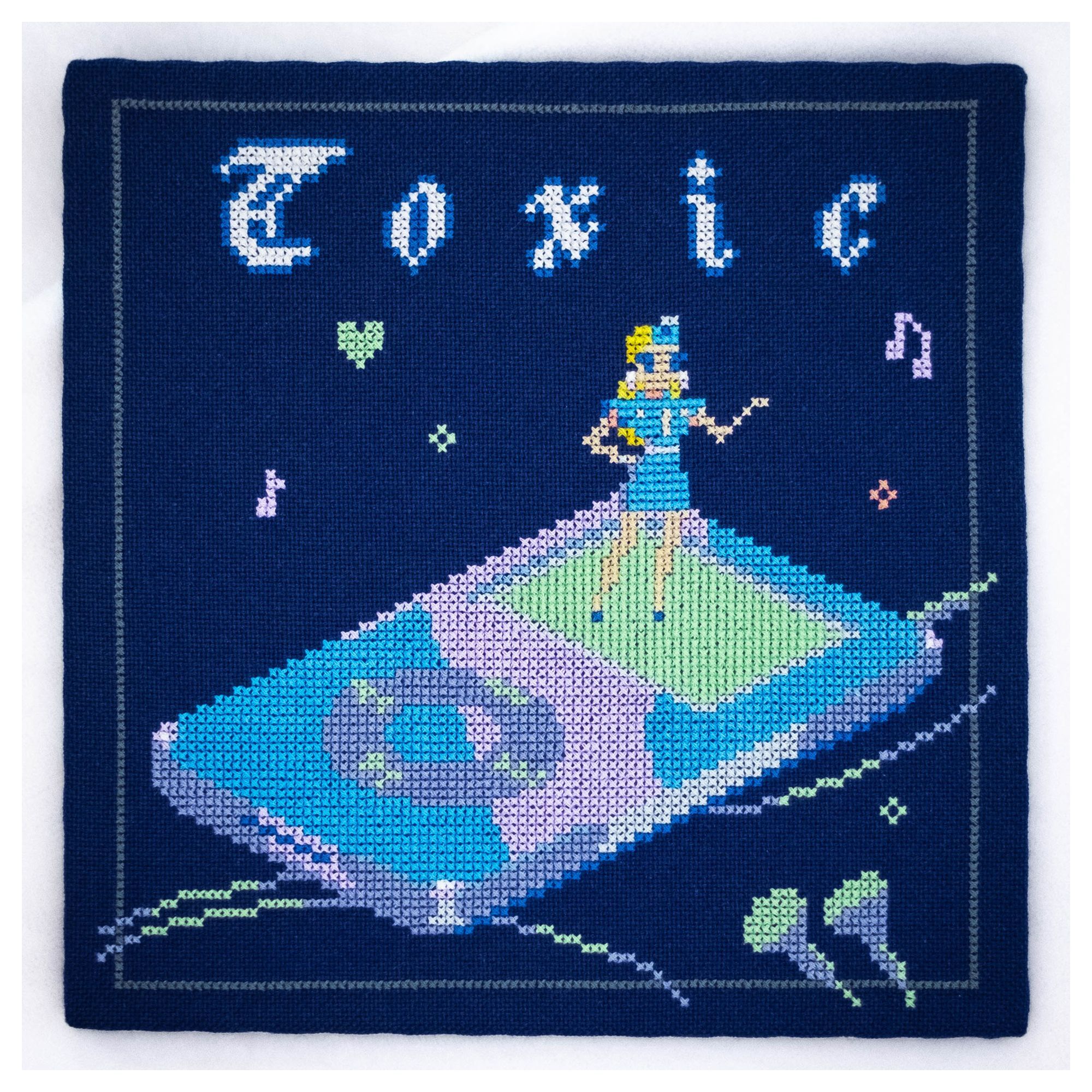 A finished cross stitch project on a dark blue fabric. Britney Spears from the Toxic music video is on a retro Ipod. The colors are mainly blue, lime green and a soft pink. Toxic is written in a gothic/juicy font