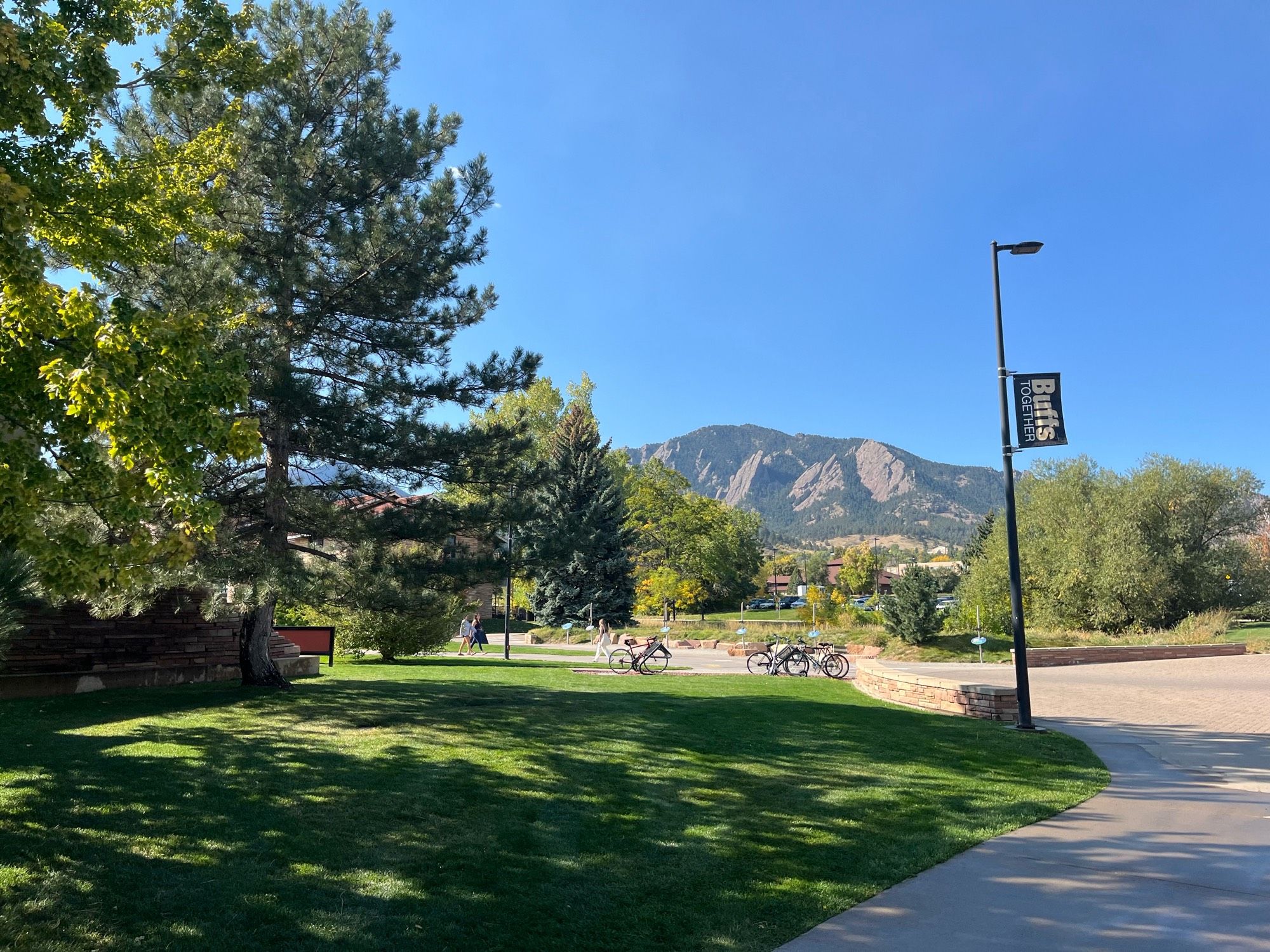 A photo taken on the campus of CU Boulder. A sidewalk, bike path, and green space dominate the foreground, while trees rise from the ground to frame the mountains and third through fifth Flatirons in the background