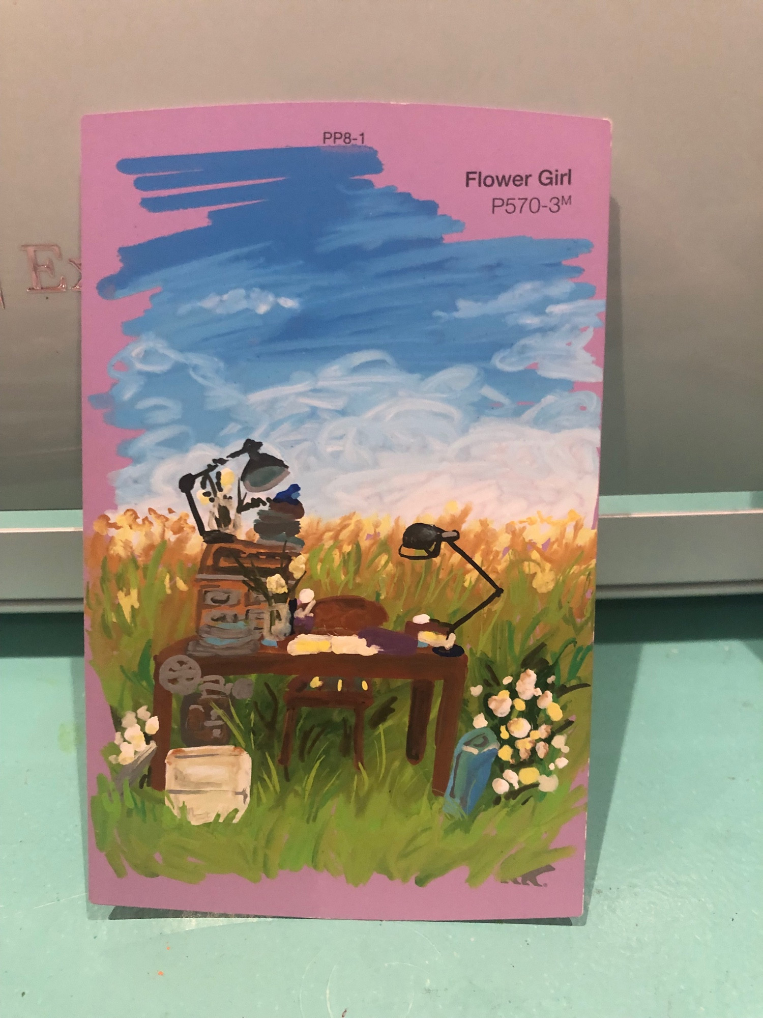 A drawing on a paint chip. The paint chips color name is Flower girl, which is a medium pinkish purple color. There’s a rectangular vertical drawing with a scribbly finish on the edges showing the pink chip color underneath. The drawing is of a grassy field with yellow grass in the distance. In the grass there’s a desk, with various objects on it, and a pot of flowers next to the desk. There’s a bright blue sky with white clouds in the back too. 