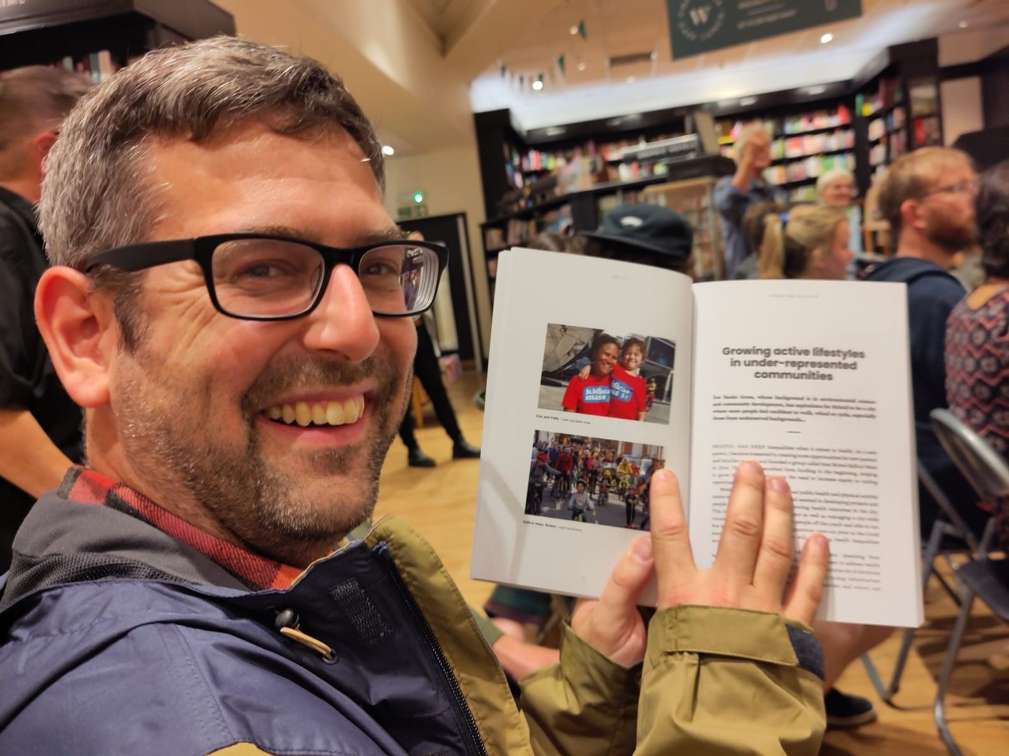 Me grinning while pointing at my picture in the book while seated waiting for a book launch event to start in Waterstones Bristol.