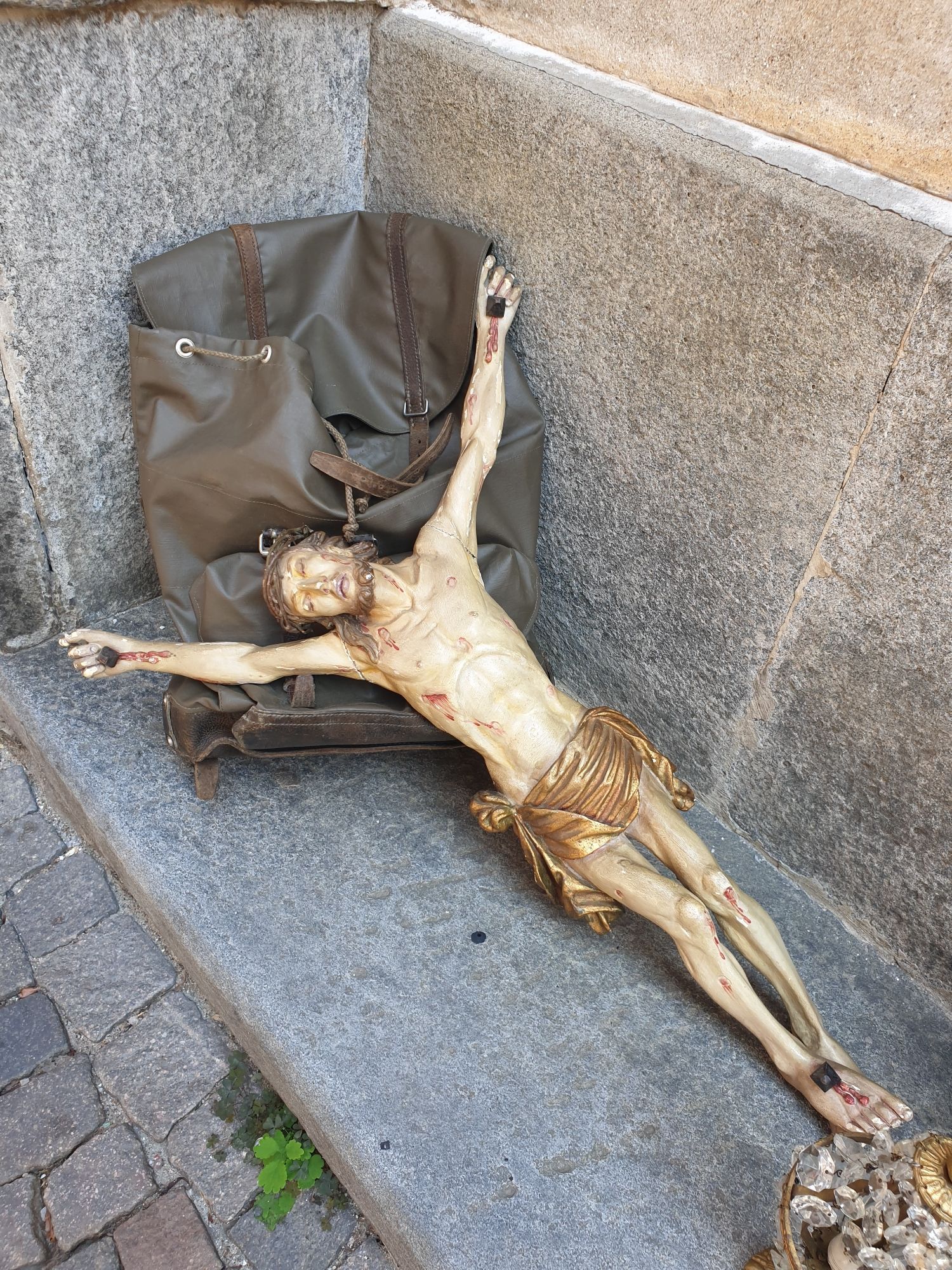 Jesus der Gekreuzigte am Flohmarkt in einer Nische, mit dem Kopf auf einem alten Militärrucksack liegend.