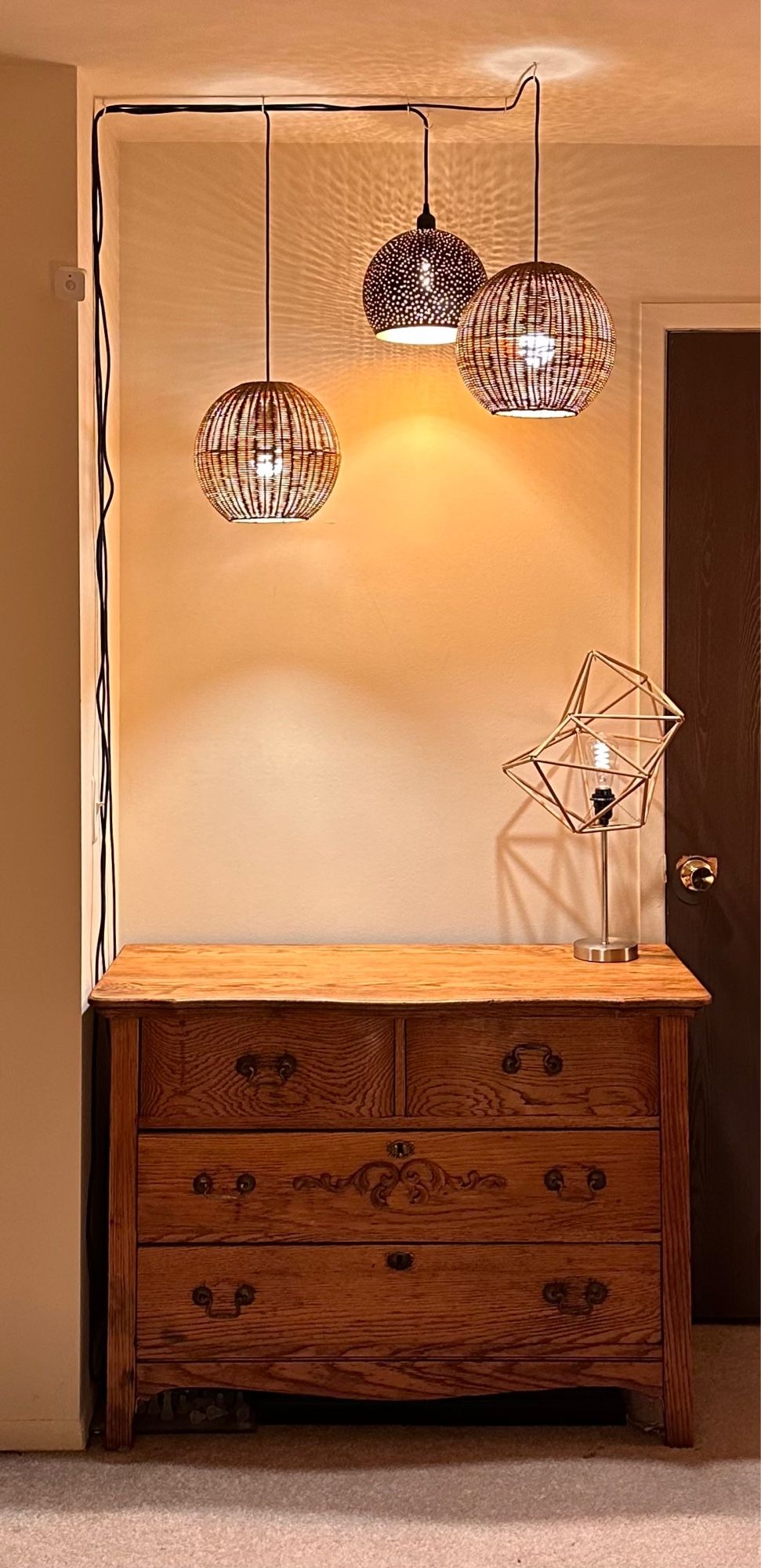 An excessive amount of lamps and an antique dresser in the corner of a white walled room. Three spherical shades, two woven and one pierced metal, hang above with black cords running down the wall on the left. A geometric wooden dowel sculpture with an Edison style LED bulb pretend to be a table lamp on the right.