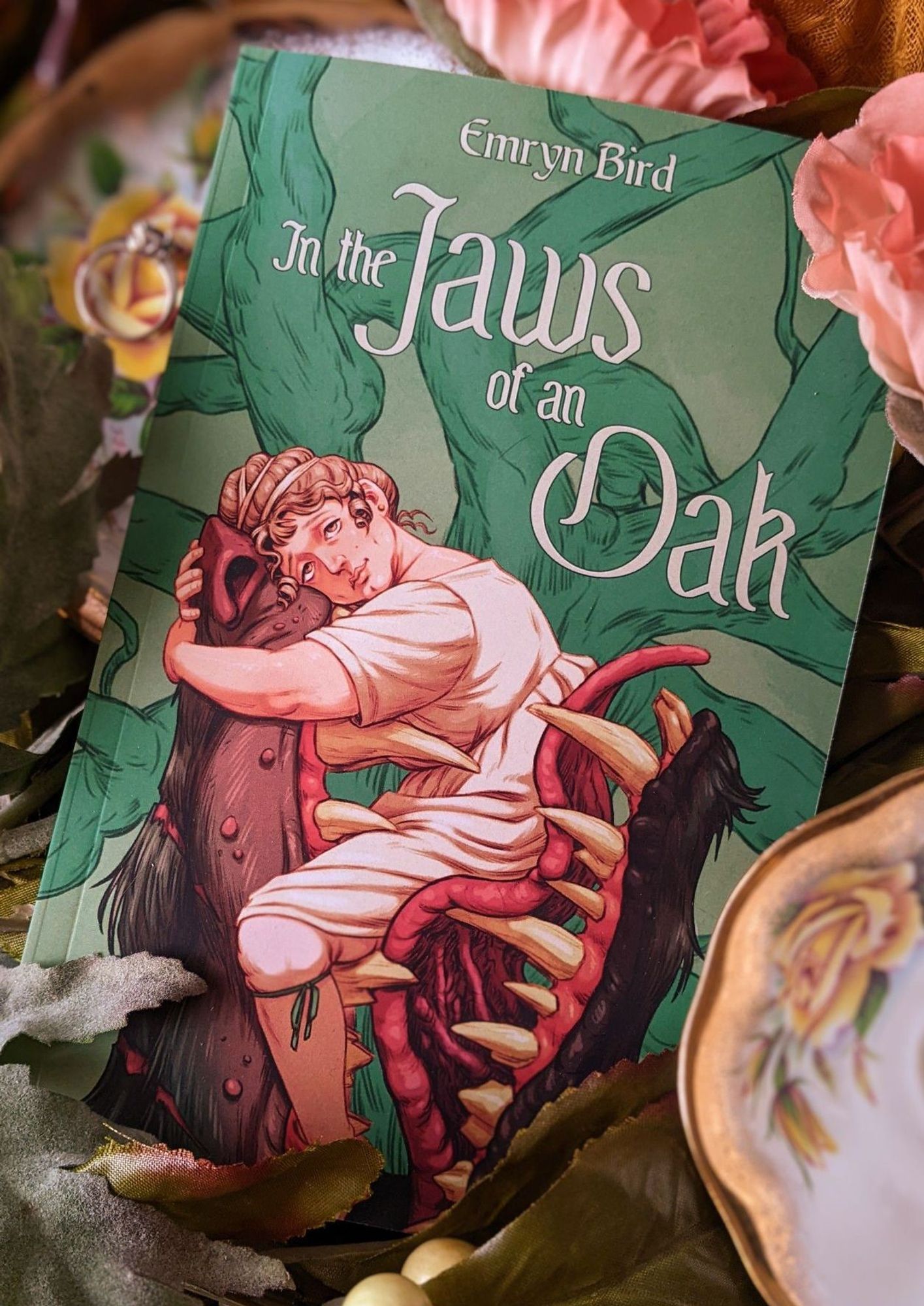 Picture of a book nestled between some fake flowers, a teacup, and saucer with a ring on it. The book In the Jaws of an Oak by Emryn Bird features a pale woman in a chemise, stockings, and teal ribbon garters sitting in the mouth of a large wolf-like beast.