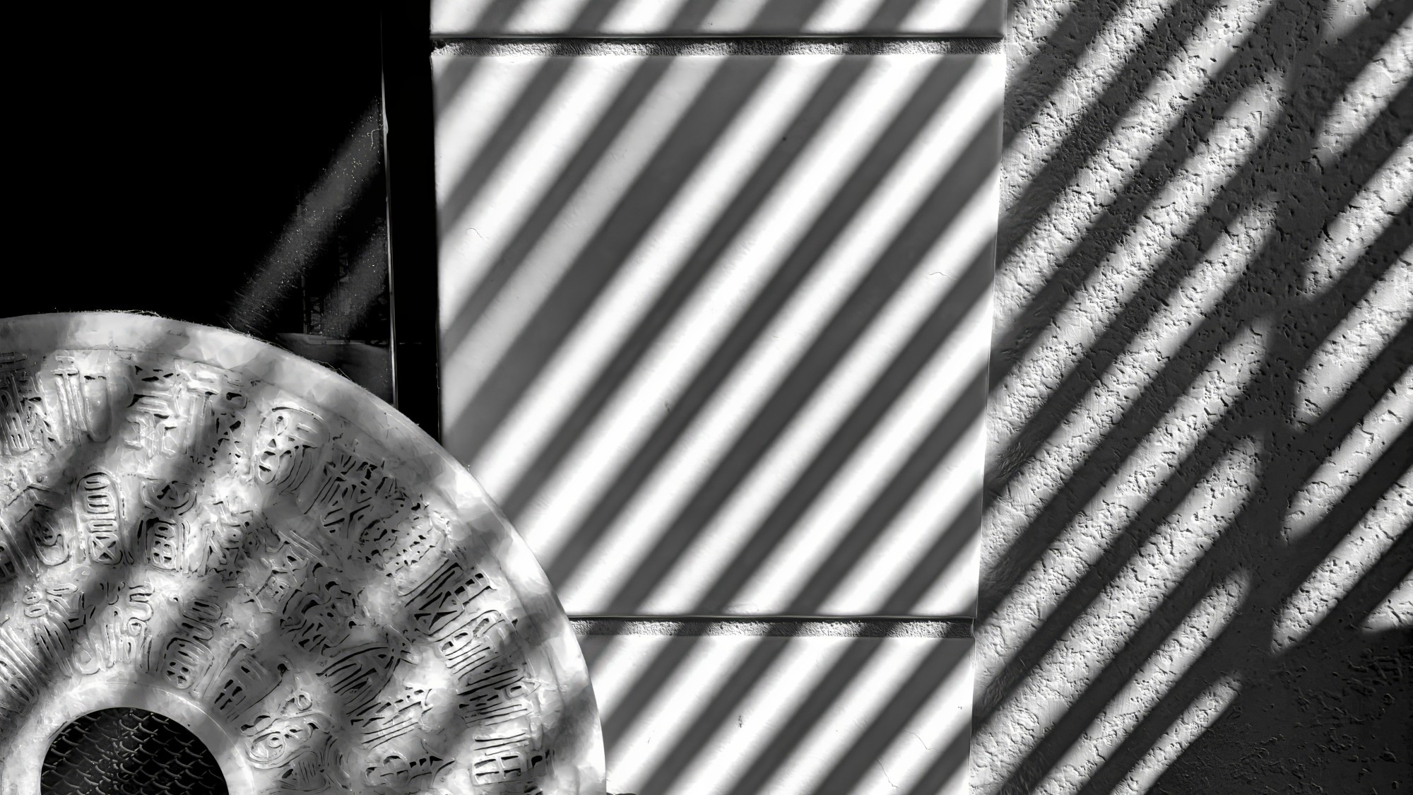 A monochrome image of the pattern of lines created by the shadow of window blinds cast on a wall and the edge of a fireplace. The semicircular object in the lower is a decorative ceramic disk decorated with some sort of obscure symbols.