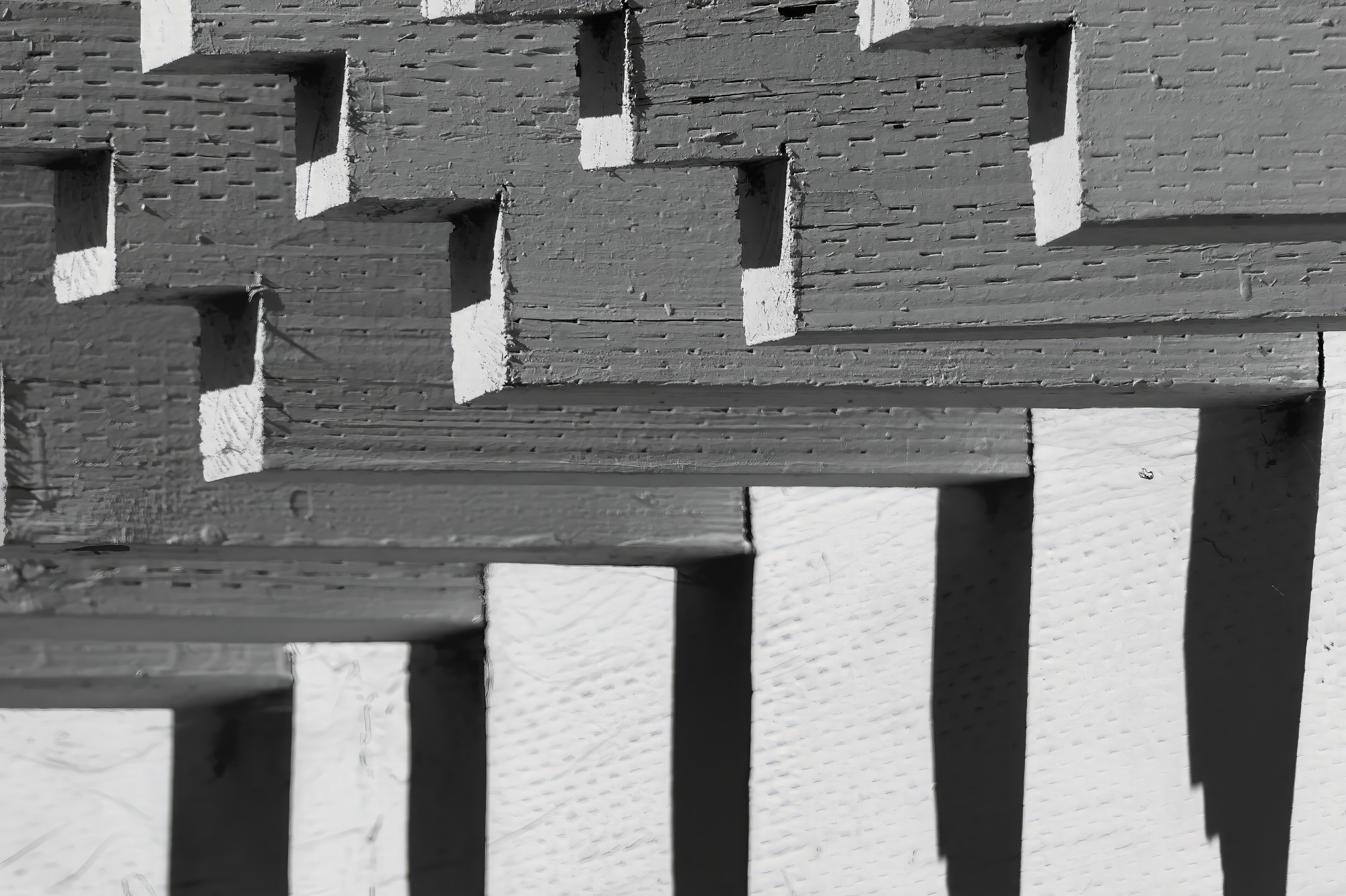 A monochrome image of the pattern of lines and shadows in a detail of some decorative woodwork on the top of a fence.