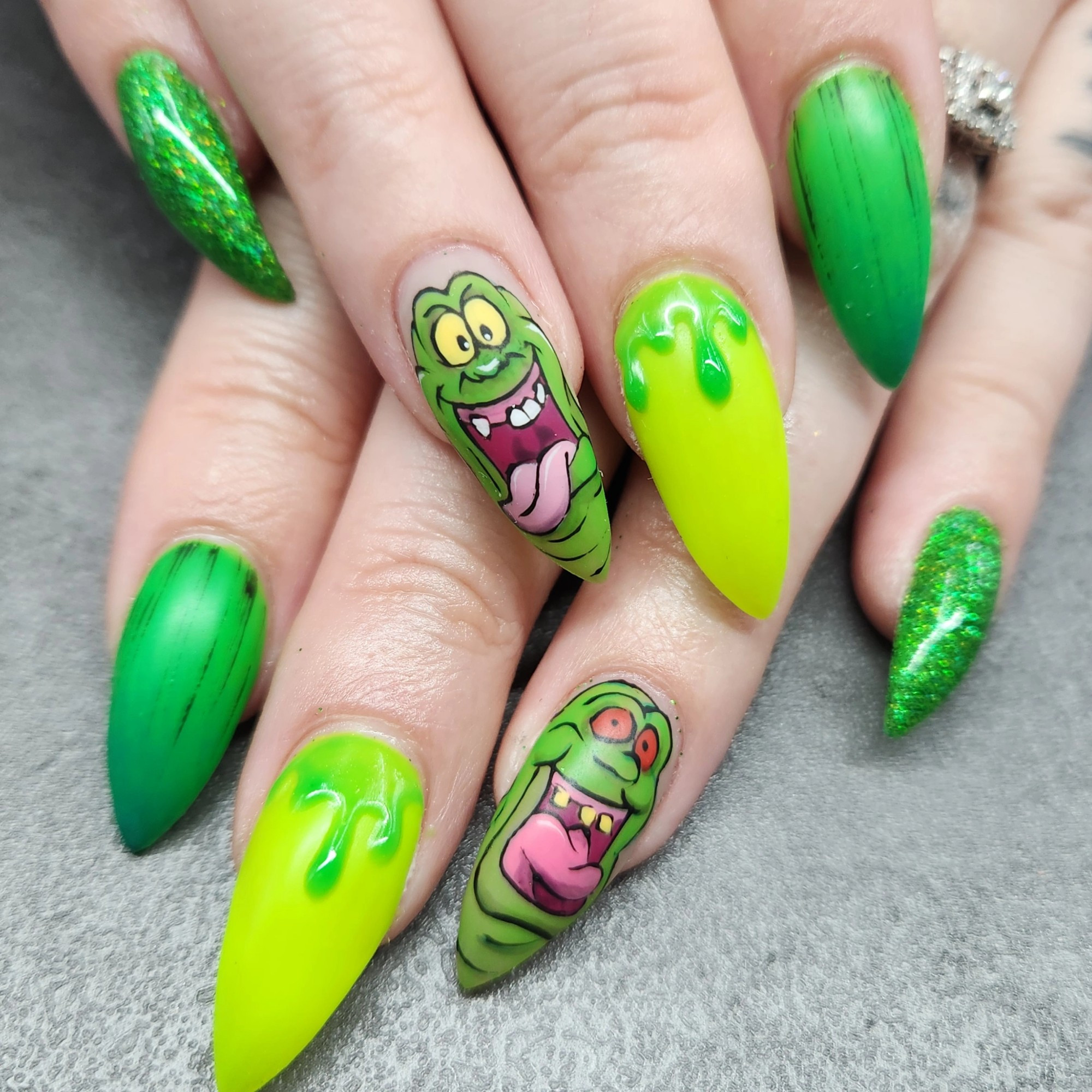 Picture of hands with long pointed nails. The nails are in shades of green with art of slime looking drips. Two of the nails have hand painted art of Slimer from Ghostbusters.