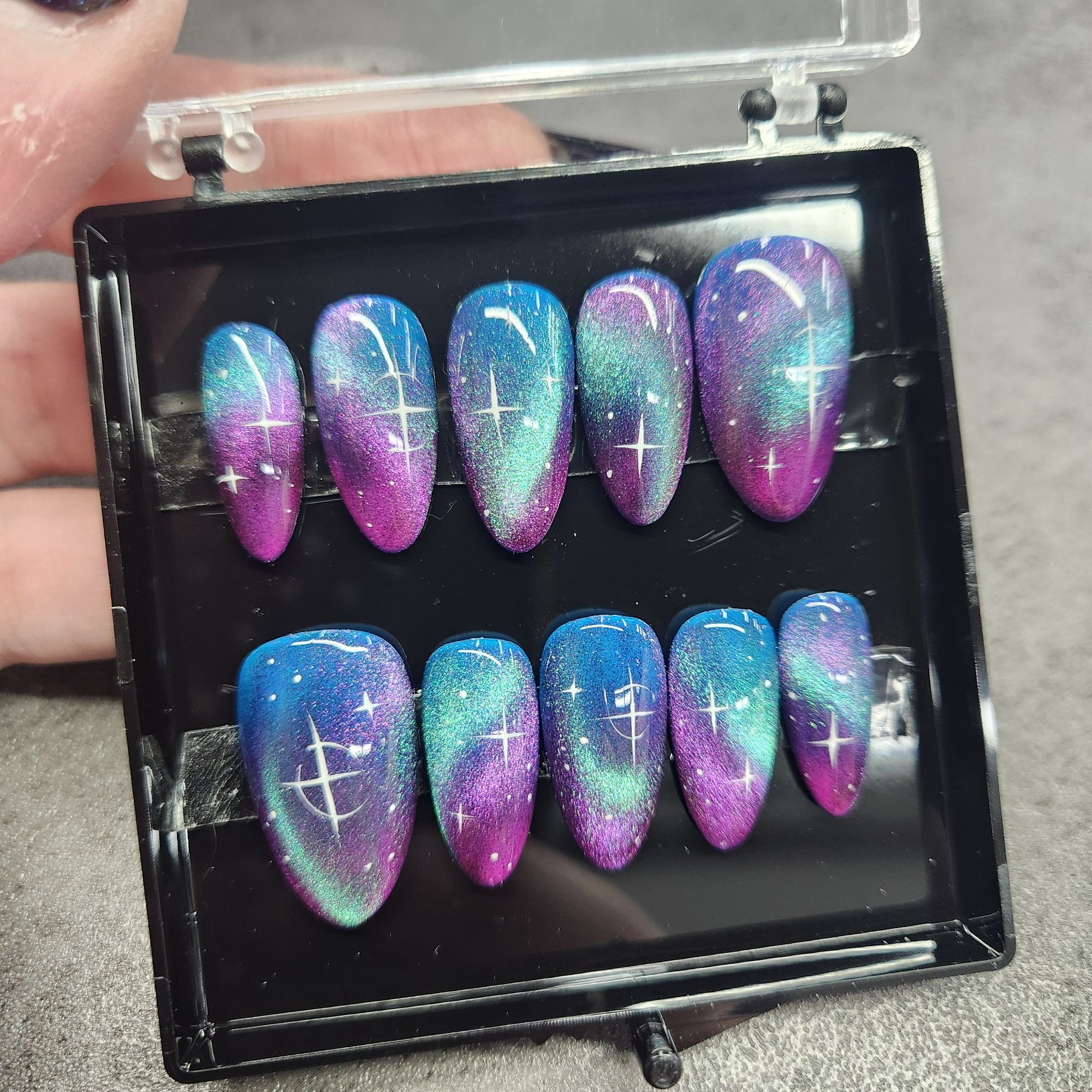 Picture of a setbof press-on nails in a black plastic container. The are painted with a galaxy design in blue, teal, and purple colors.