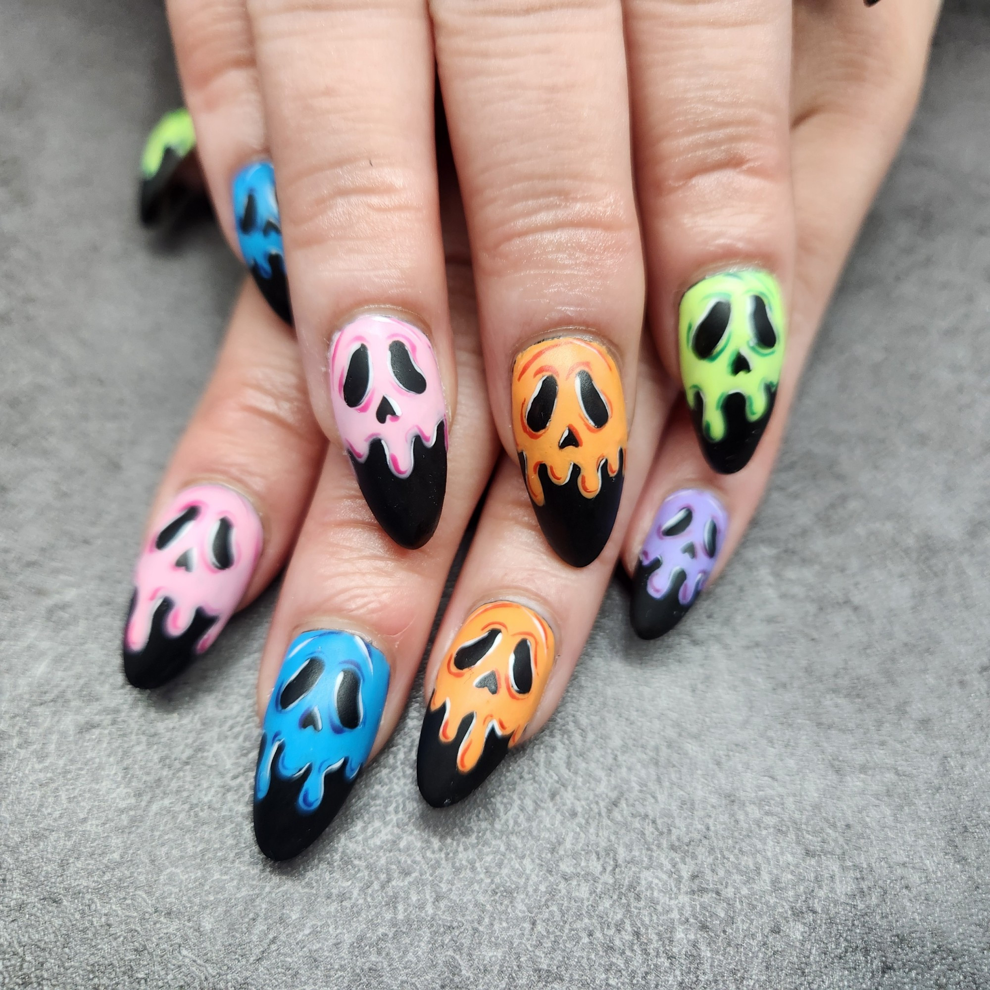 picture f hands with almond shaped nails. Nails are matte black with drippy skulls painted on them in different colors.