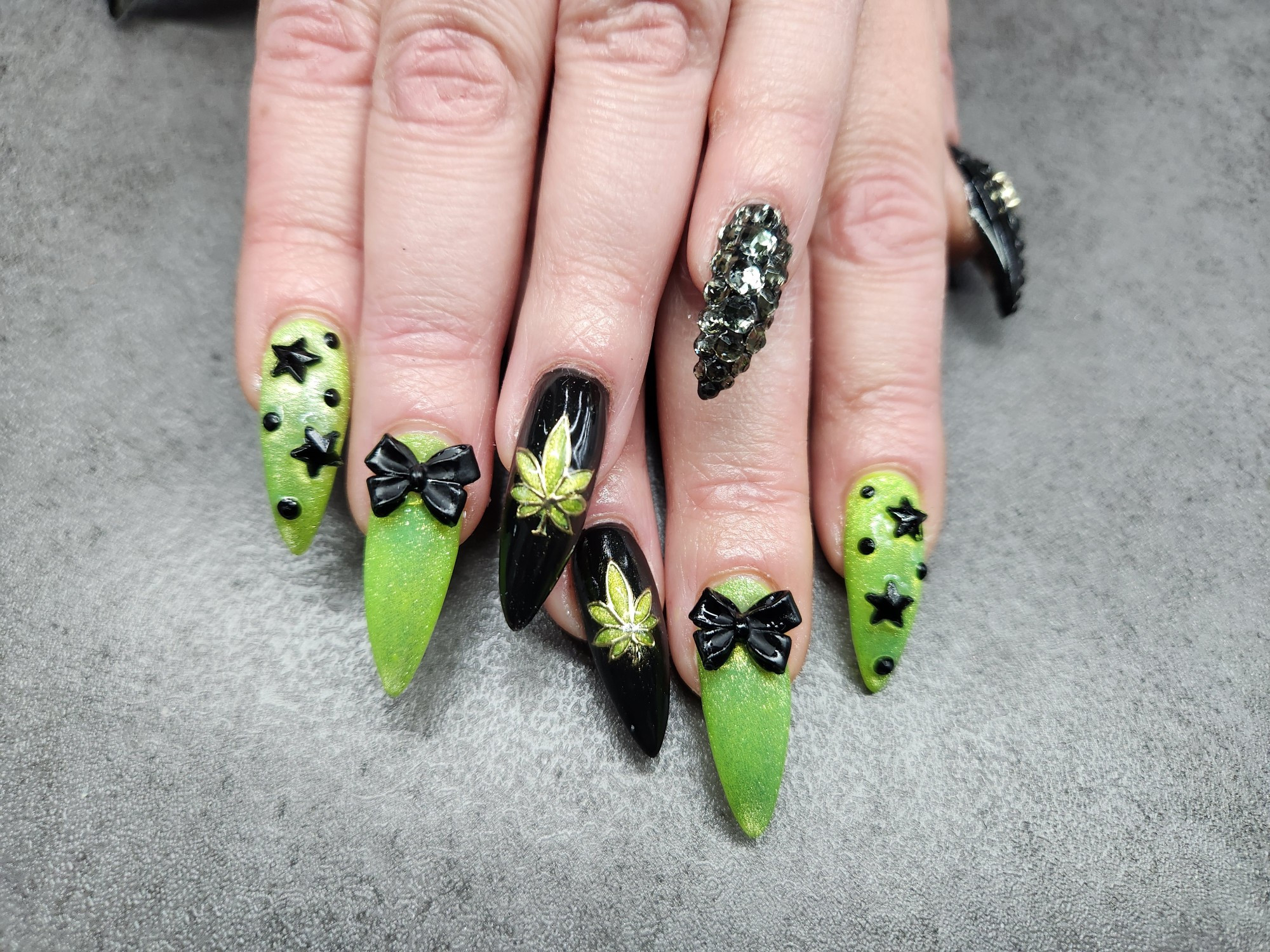 Picture of hands with long pointed nails, with Brat Summer inspired nail art. Nails are green with black gems and bows, a few nails are black with cannabis leaves painted on them.