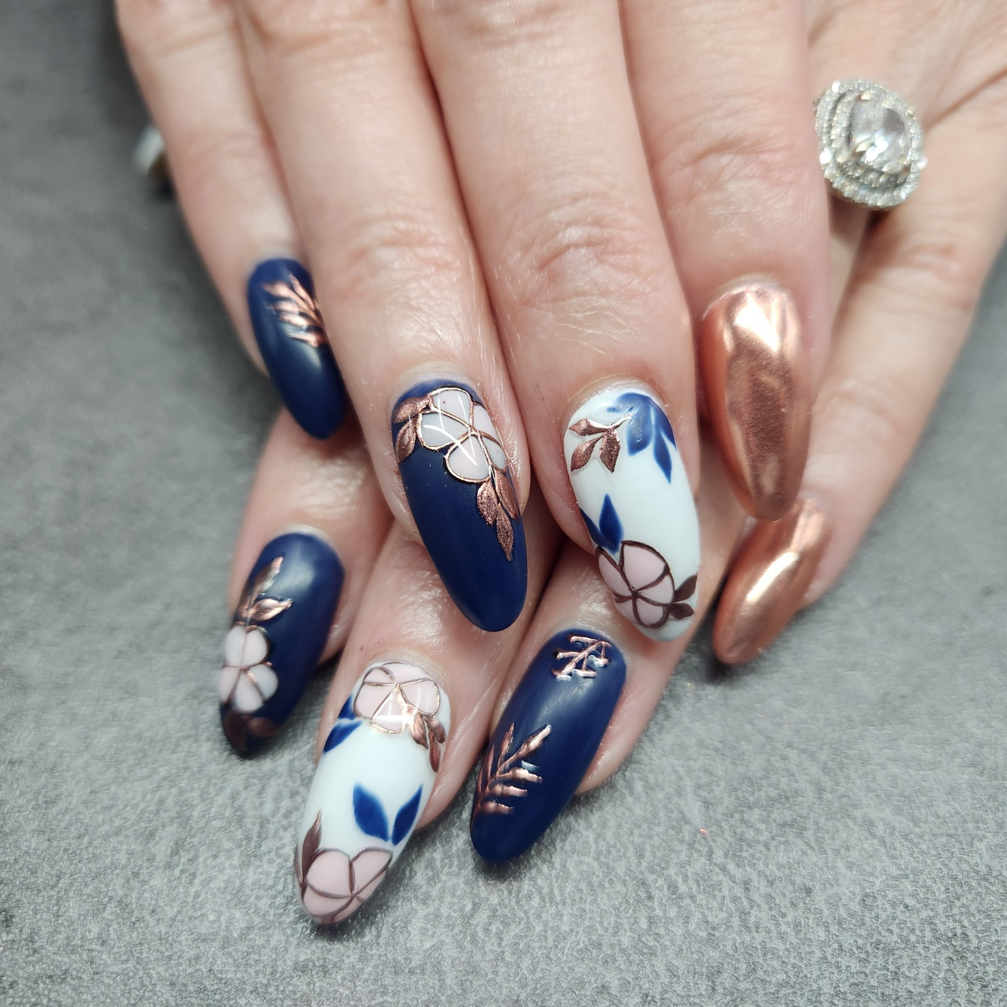 hands with long oval shaped nails. Nails are matte with porcelain inspired floral nail art in navy blue, white, and pink, with rose gold chrome accents