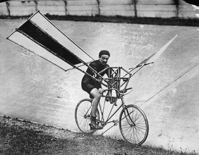 Black and white photo of a human-powered flight attempt from the late 19th or early 20th century: a man rides a bicycle to which flapping ‘wings’, likely made of light wood and fabric, have been attached.