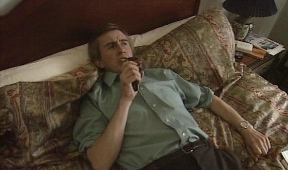 Still image from the TV series I’m Alan Partridge, showing the title character lying on a hotel bed wearing a green shirt and grey trousers, and speaking into a dictaphone.